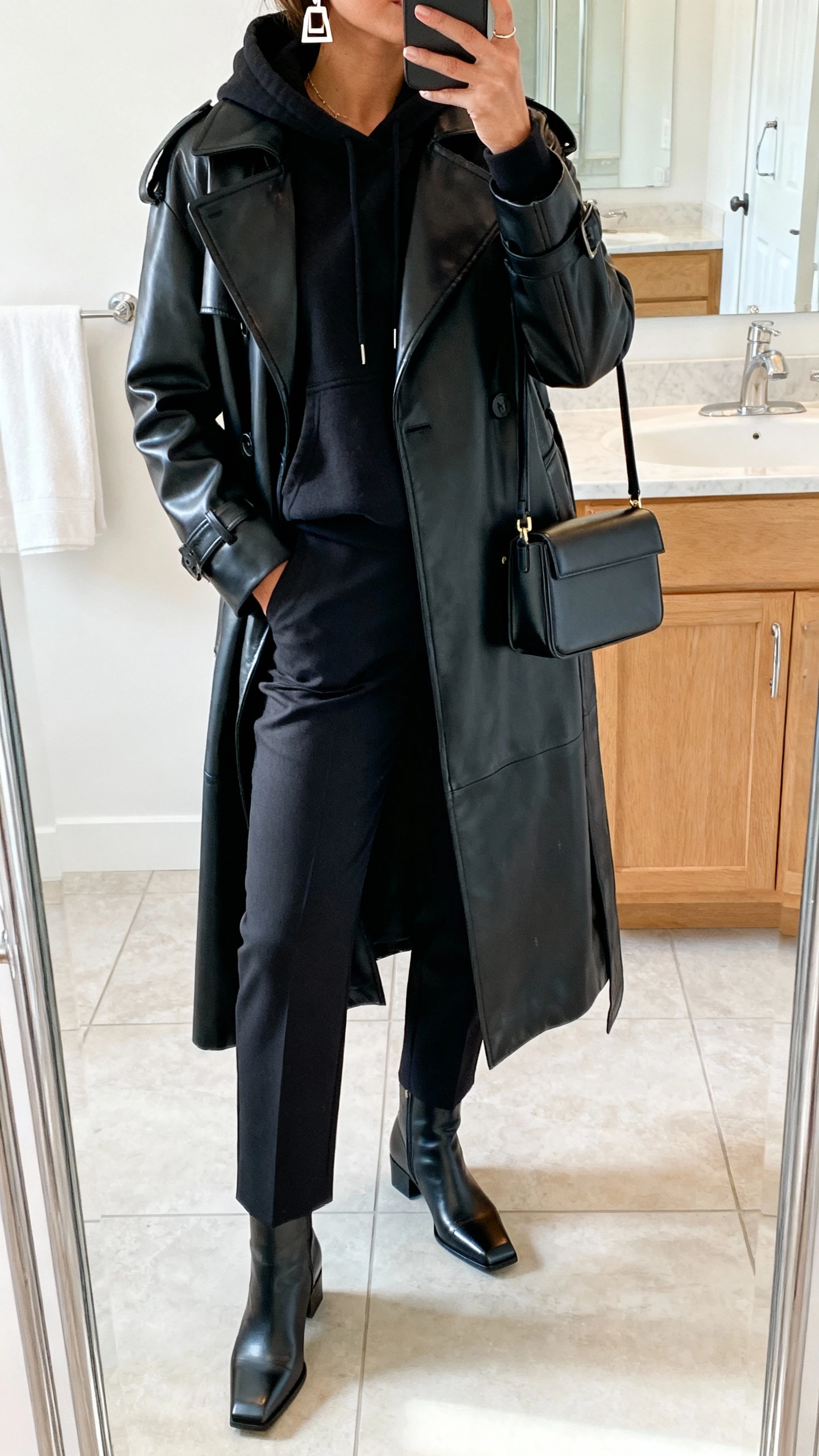 Bathroom mirror selfie of a woman wearing a black leather trench over a hoodie, tailored trousers, square-toe boots, structured mini bag, geometric earrings, face not visible, cropped at neck, bright indoor lighting, natural iPhone photo.