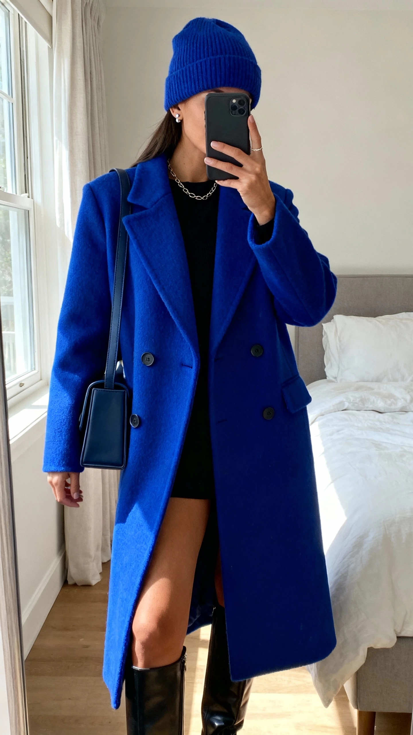 Bedroom mirror selfie of a woman in a vivid single-breasted wool coat (cobalt) over a black base layer, knee-high boots, boxy shoulder bag, cashmere beanie in matching tone, simple studs and delicate chain, face not visible, cropped at neck, morning window light, natural iPhone quality.