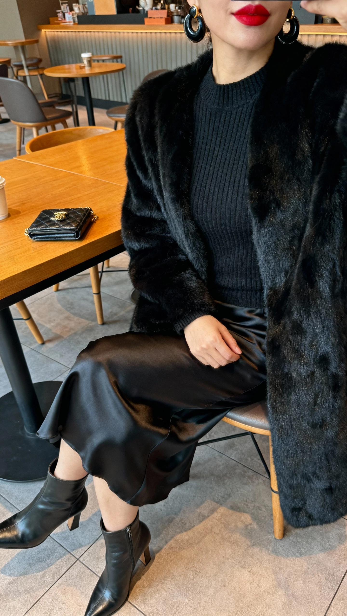 Cafe table side-angle selfie of a woman in a jet black faux fur coat over a fitted knit, satin slip skirt, heeled boots, mini clutch, bold earrings and red lip implied (face not visible), cropped at chin down, warm cafe lighting, natural iPhone quality.