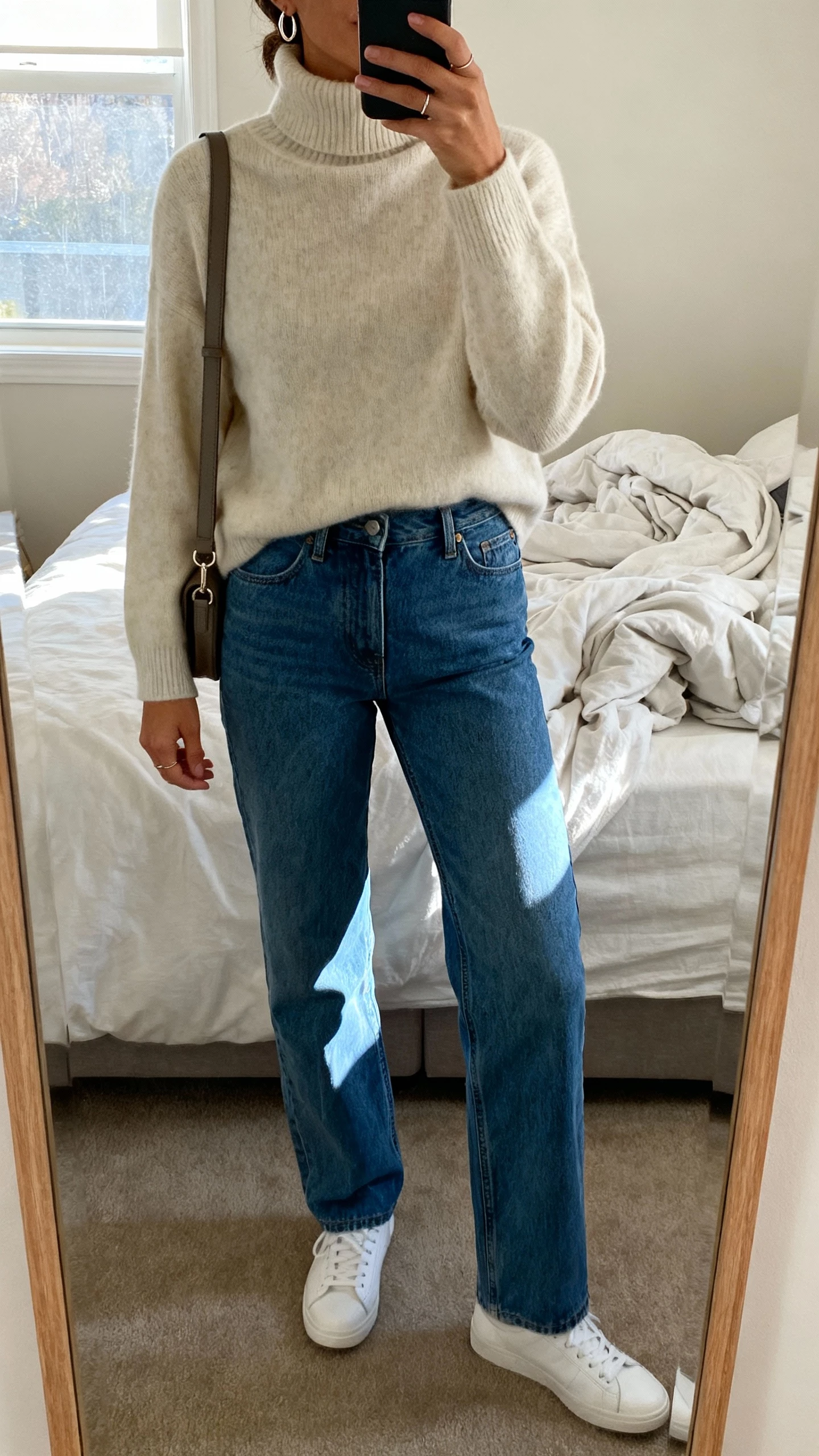 Casual iPhone mirror selfie of a woman in a cashmere turtleneck with straight-leg blue denim, white sneakers, crossbody bag, delicate hoops, face not visible, bedroom mirror with unmade bed, soft afternoon window light, natural iPhone photo quality.