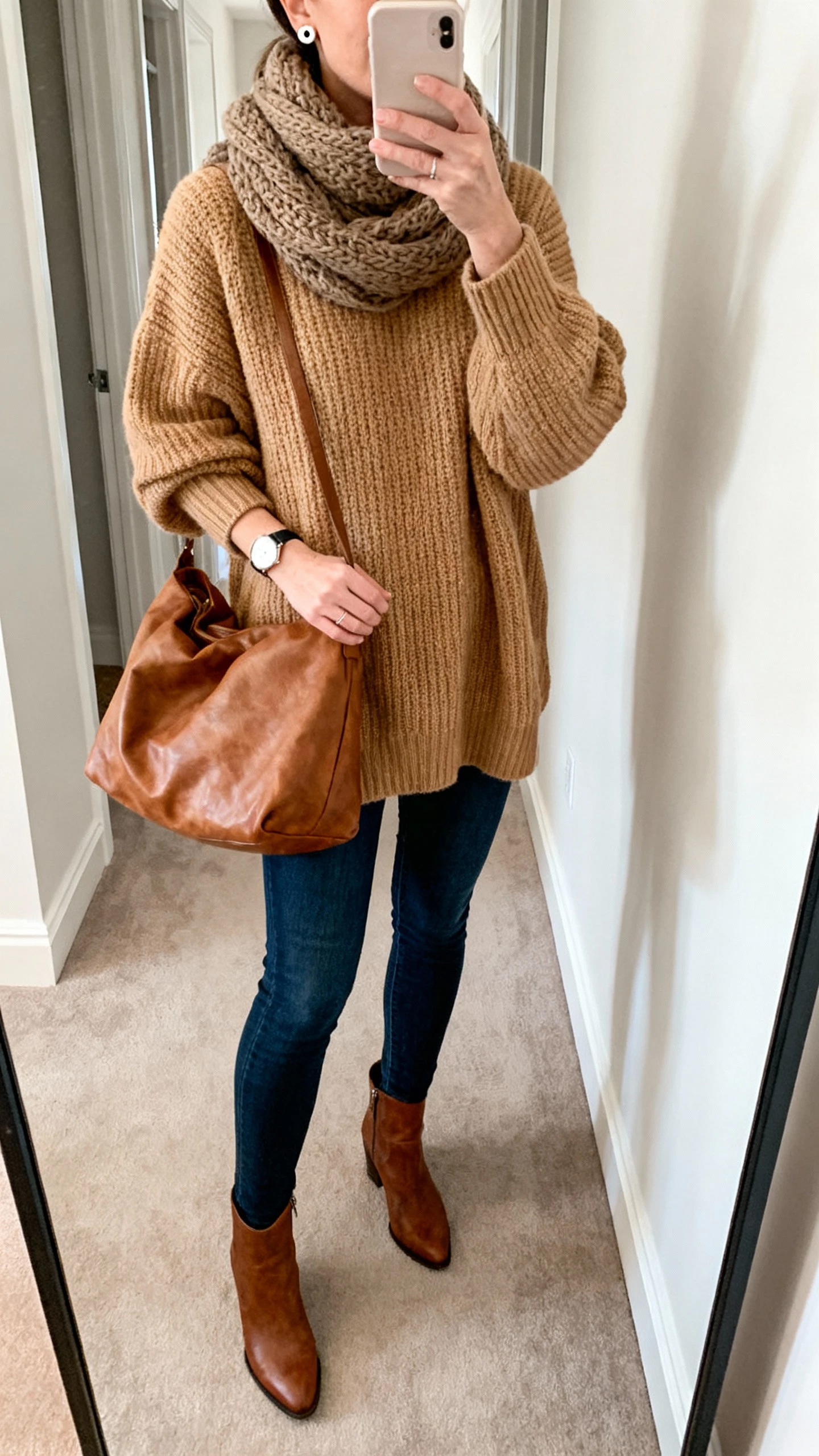 Casual iPhone mirror selfie of a woman in an oversized camel knit sweater with dark blue skinny jeans, leather ankle boots, chunky scarf, stud earrings, simple watch, and a slouchy leather hobo bag; cropped at shoulders, hallway mirror, soft morning light, face not visible, natural iPhone aesthetic.