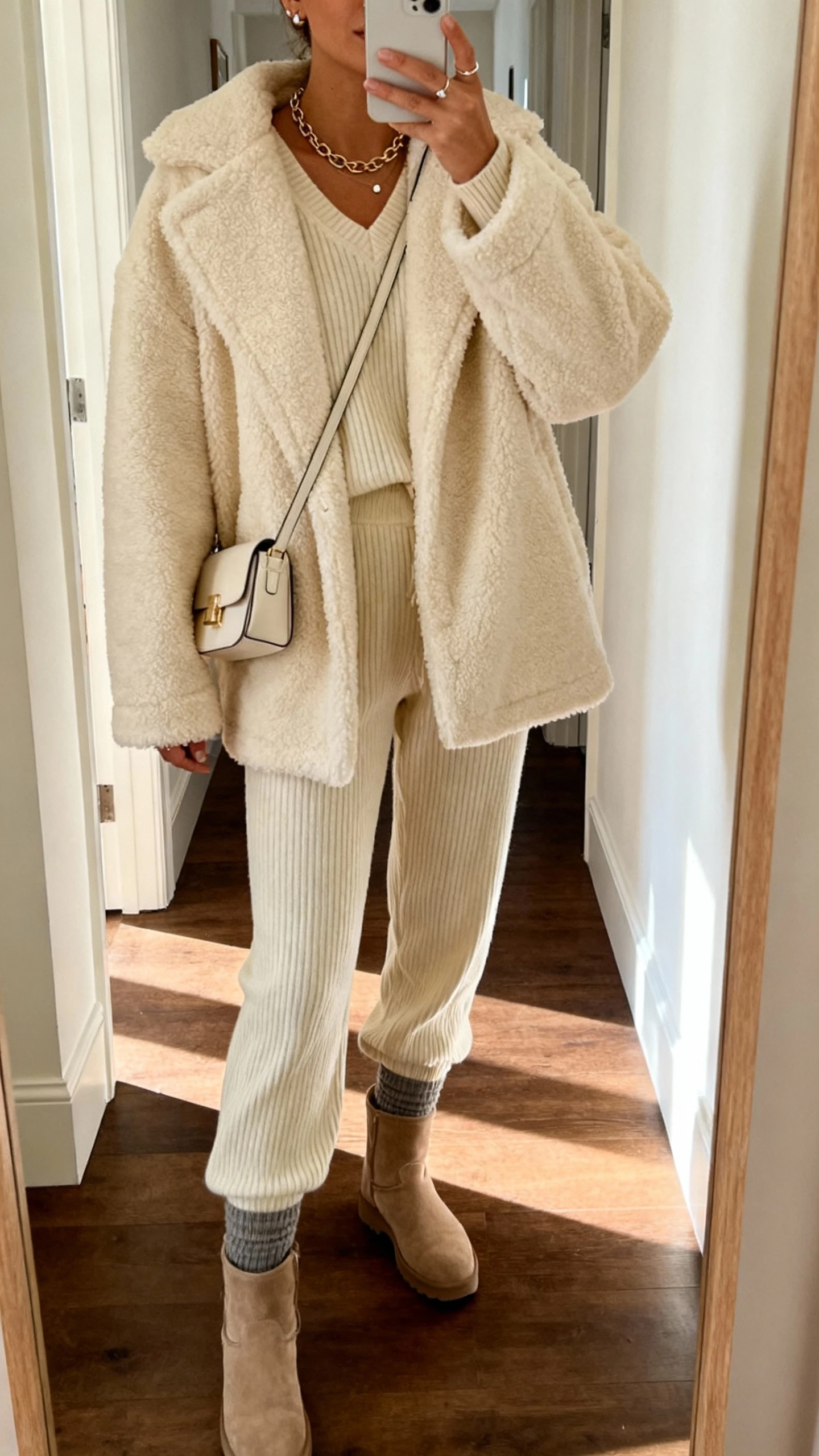 Cozy hallway mirror selfie of a woman wearing a cream teddy coat over a ribbed knit set (sweater and pants), sock boots, mini shoulder bag, delicate chain necklace and studs, face not visible, cropped at shoulders, warm indoor lighting, natural iPhone quality.