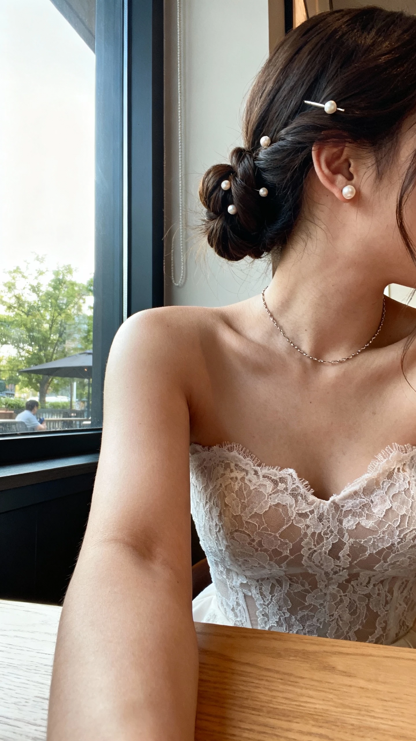 Cropped-at-shoulders iPhone selfie of a woman in a lace bodice dress, half-up twist secured with small pearl pins, dainty pearl studs and a thin chain, face not visible, sitting at a cafe table before the ceremony, morning window light, natural iPhone aesthetic.
