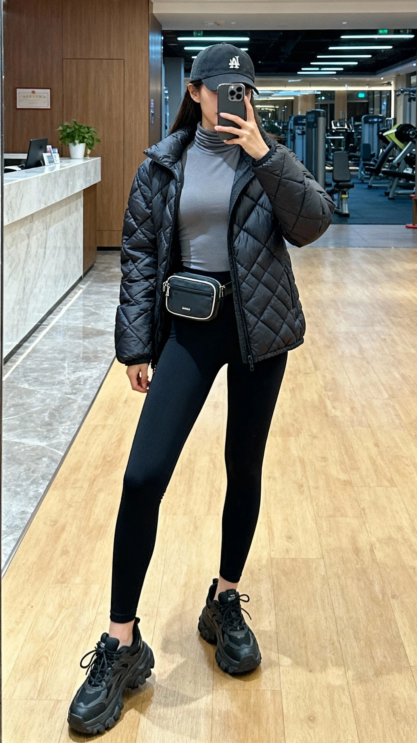 Gym-lobby iPhone mirror selfie of a woman wearing a fitted turtleneck under a quilted puffer, black leggings, chunky sneakers, baseball cap, mini belt bag, face not visible, indoor lighting, casual iPhone photo.