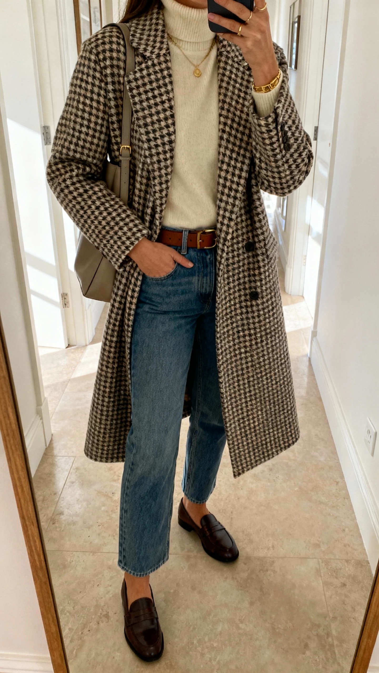 Hall mirror selfie of a woman in a houndstooth checked wool coat, cream cashmere turtleneck, straight-leg denim, classic loafers, leather belt, medium shoulder bag, subtle gold jewelry, face not visible, cropped at shoulders, indoor daylight, natural iPhone quality.
