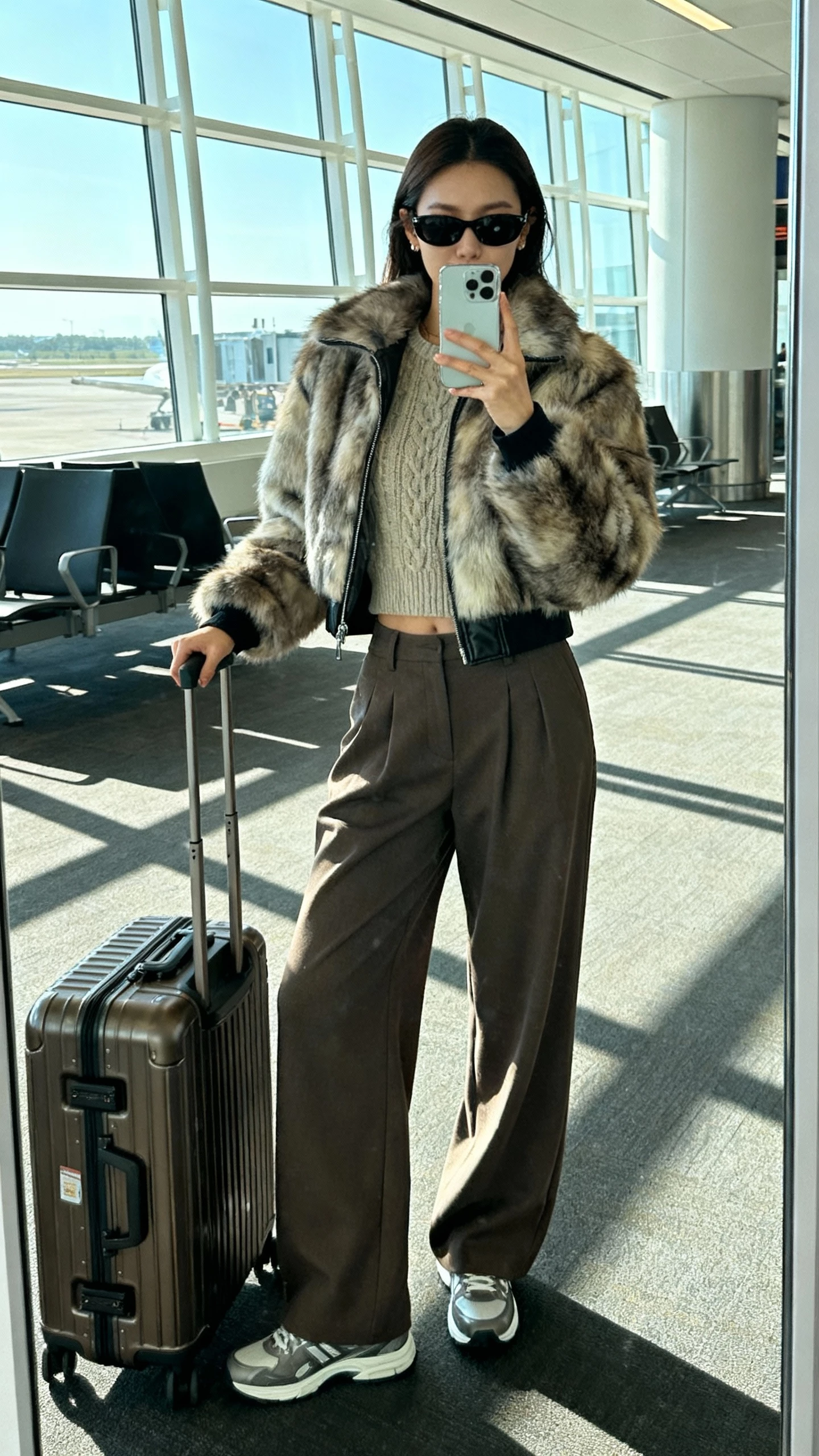 iPhone airport mirror selfie of a woman wearing a fur bomber jacket, wide-leg trousers, sleek sneakers, structured carry-on, knit top, sunglasses, face not visible, terminal lighting with window daylight, natural iPhone photo quality.