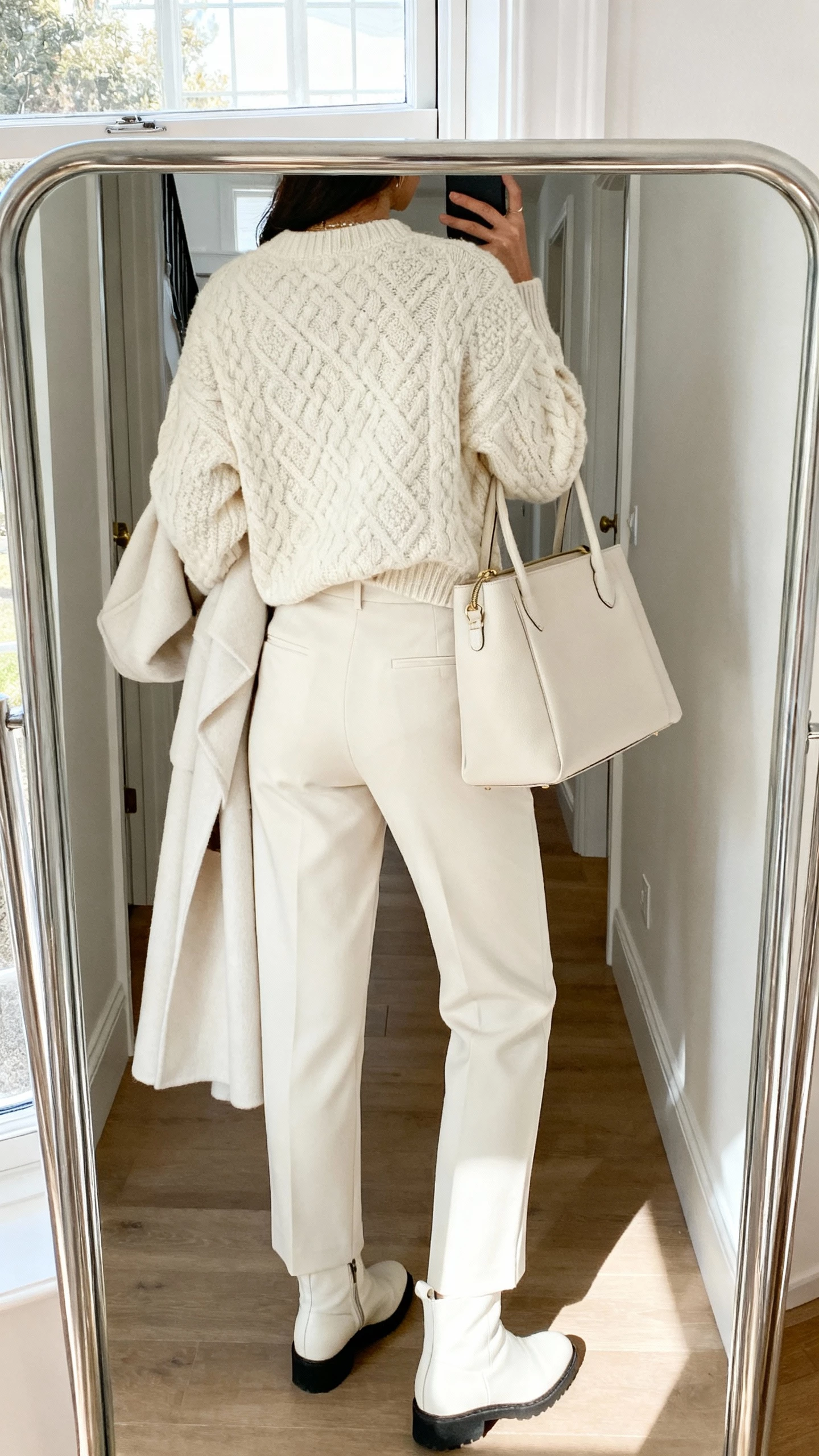 iPhone back-view hallway mirror selfie of a woman in monochrome winter whites: cream textured sweater, ivory tailored trousers, off-white coat, tonal handbag, and white boots—face not visible, entryway mirror, daylight from window, natural iPhone photo quality.
