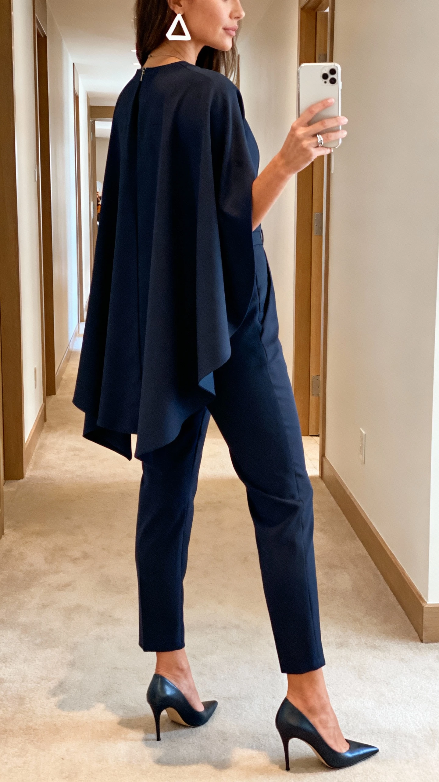 iPhone back-view hallway selfie of a woman in a tailored jumpsuit with a cape overlay, sleek pointed heels, geometric earrings, minimalist ring, face not visible, event venue corridor, soft indoor lighting, natural iPhone photo quality