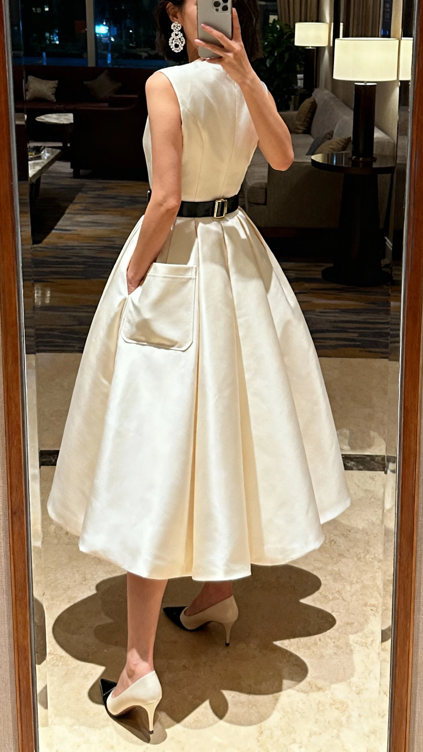 iPhone back-view mirror selfie of a woman in a modern ballgown with pockets (hands tucked in), statement earrings, sleek belt, pointed pumps, face not visible, hotel lobby mirror, evening indoor lighting, natural iPhone photo quality.