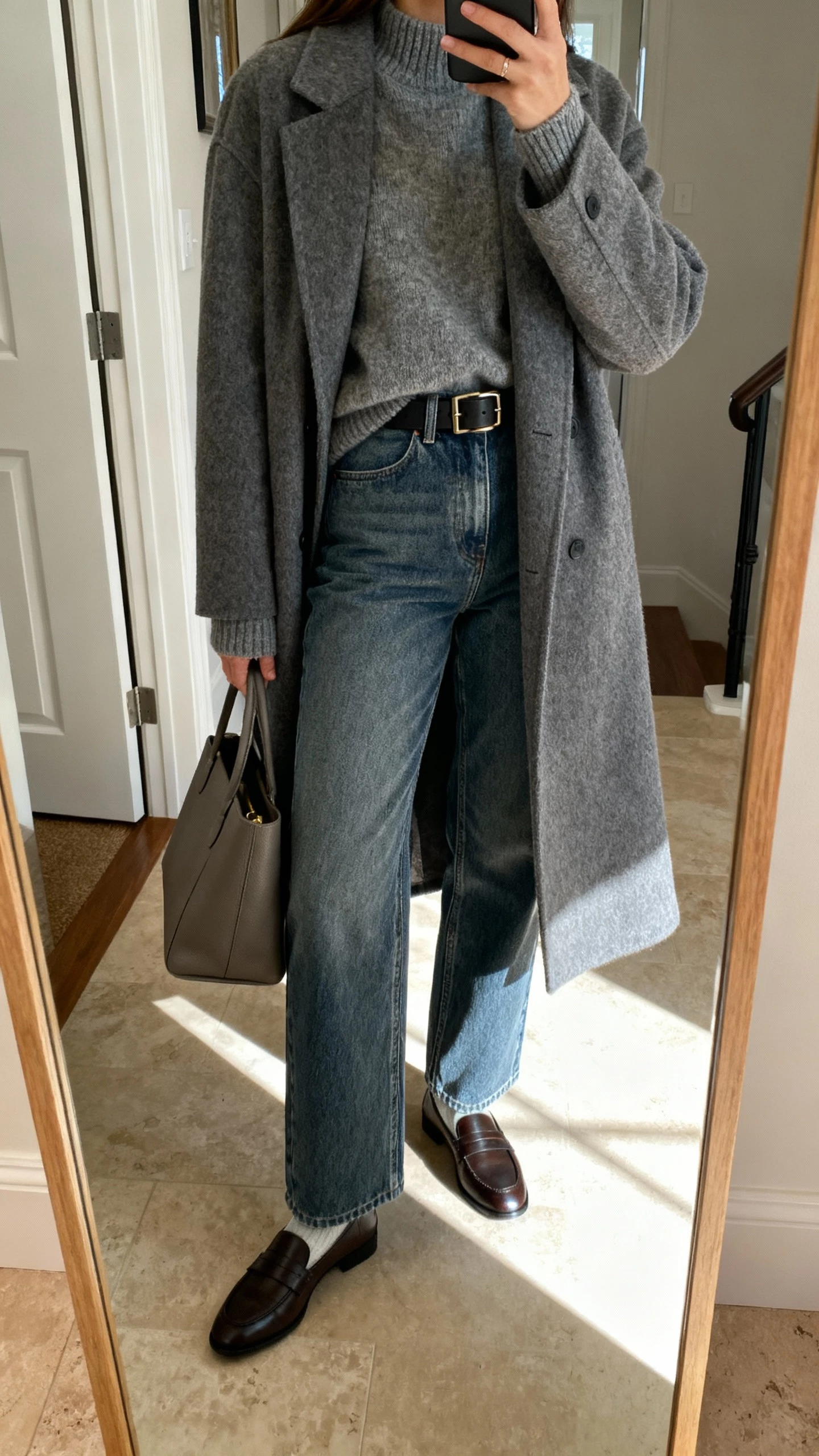 iPhone back-view mirror selfie of a woman in a monochrome wool coat over elevated straight-leg denim, a tonal sweater, loafers, structured tote, and a belt, face not visible, entryway full-length mirror with daylight, natural iPhone photo quality.