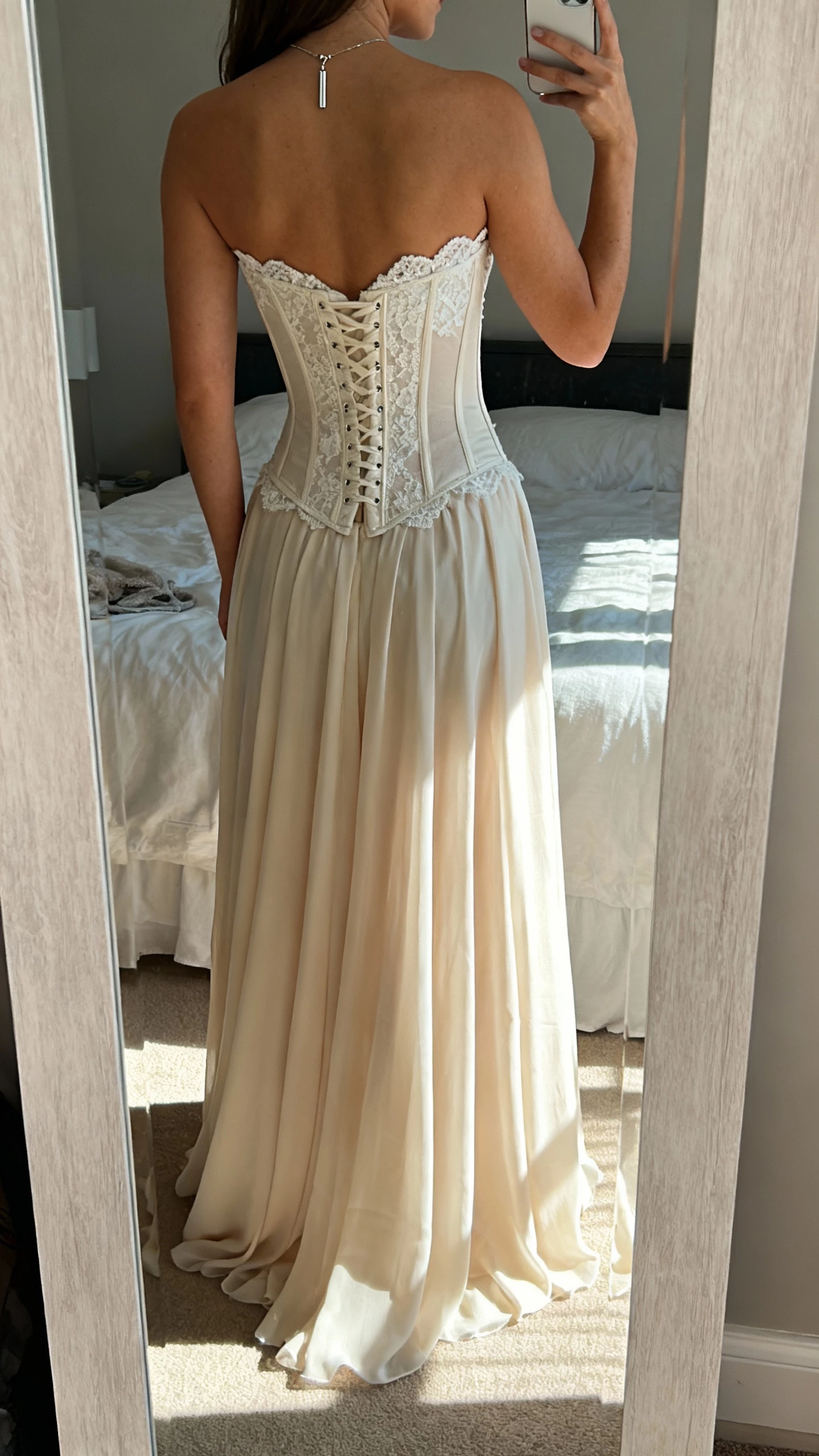 iPhone back-view mirror selfie of a woman in a Studio Levana gown with built-in corsetry for bust support, sweetheart neckline, lace bodice, and flowing chiffon skirt, accessorized with a simple pendant; face not visible, bedroom mirror, natural daylight, iPhone photo.
