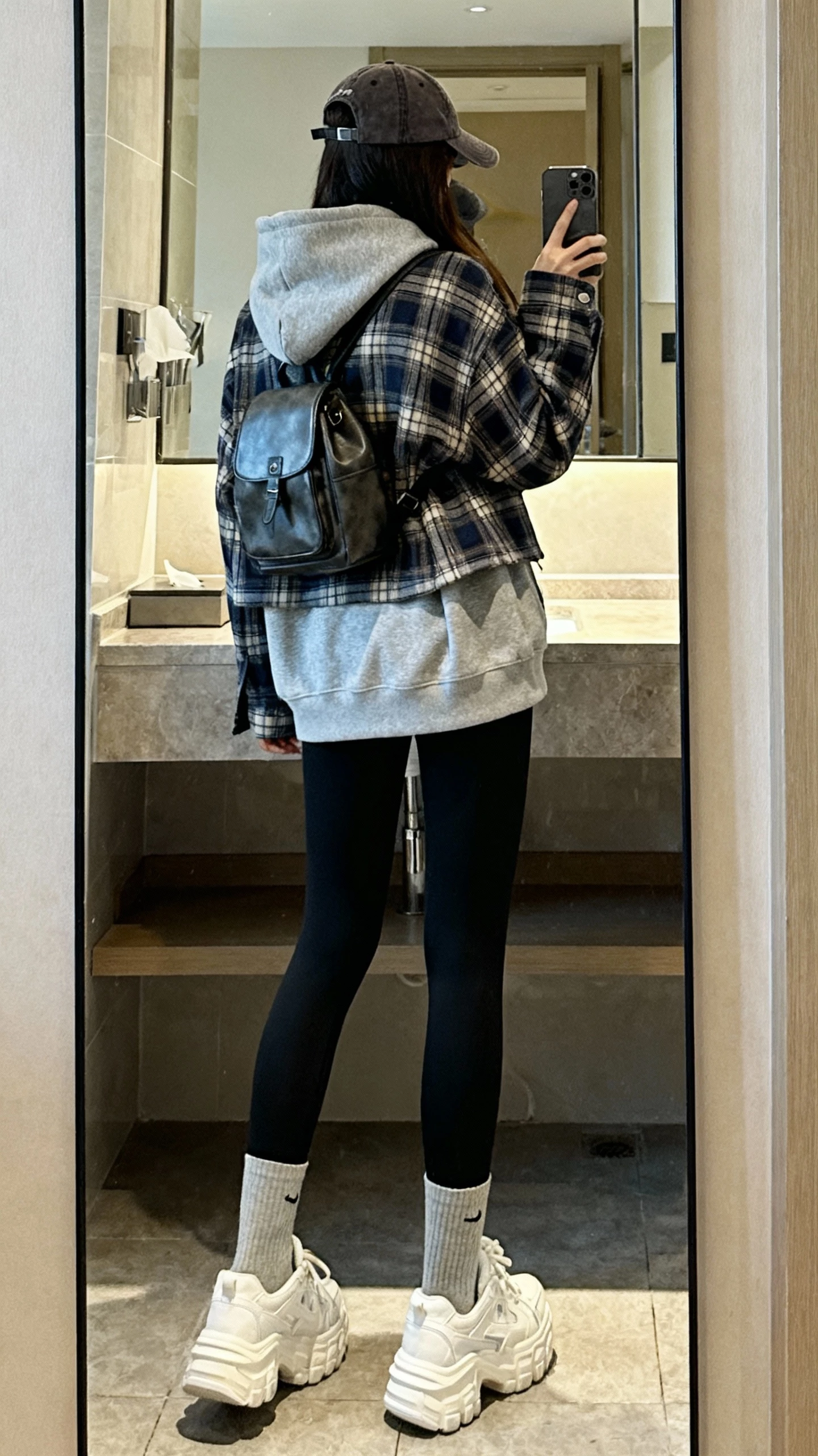iPhone back-view mirror selfie of a woman wearing a plaid shacket layered over a heather hoodie with sleek black leggings, chunky socks with clean white sneakers, baseball cap and small backpack, face not visible, bathroom mirror, soft indoor lighting, casual iPhone photo