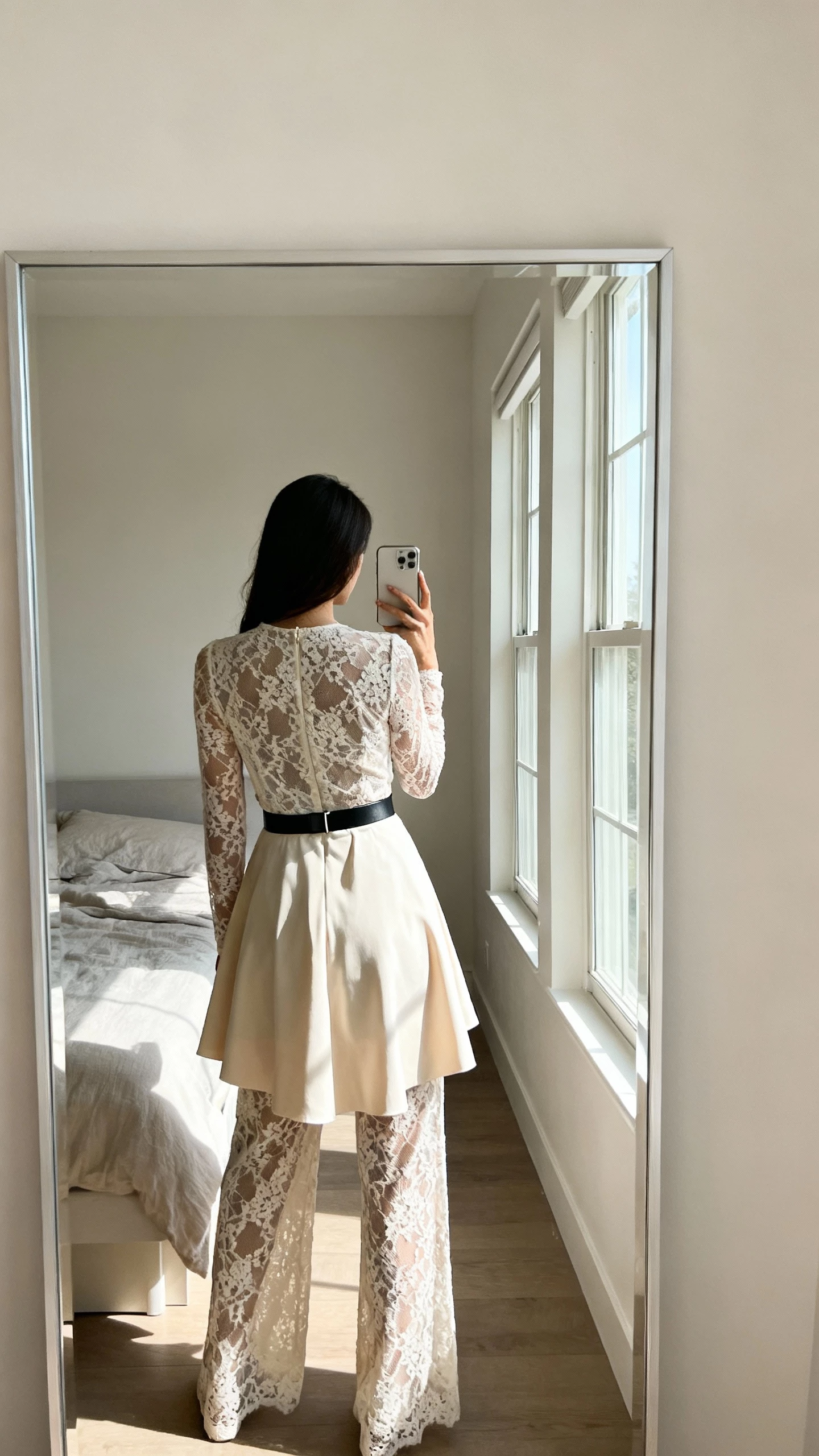 iPhone back-view mirror selfie of a woman wearing a modern lace jumpsuit with a detachable overskirt, sleek belt detail, face not visible, minimalist bedroom mirror, morning window light, natural iPhone aesthetic