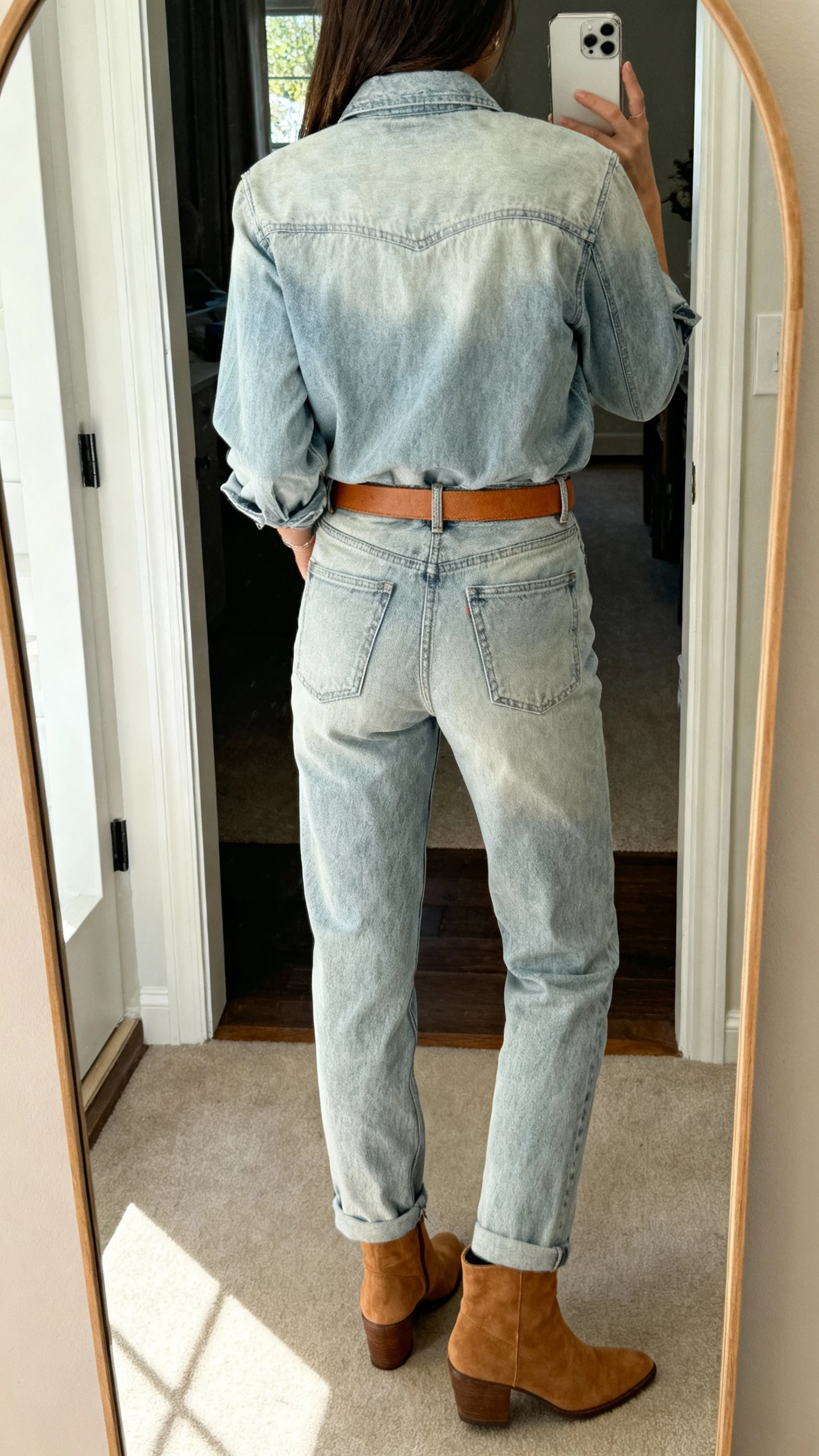 iPhone back-view selfie in a full-length mirror showing a woman wearing a light-wash denim shirt with matching light-wash jeans, tan belt, tan ankle boots, face not visible, entryway mirror, natural daylight, casual iPhone photo.