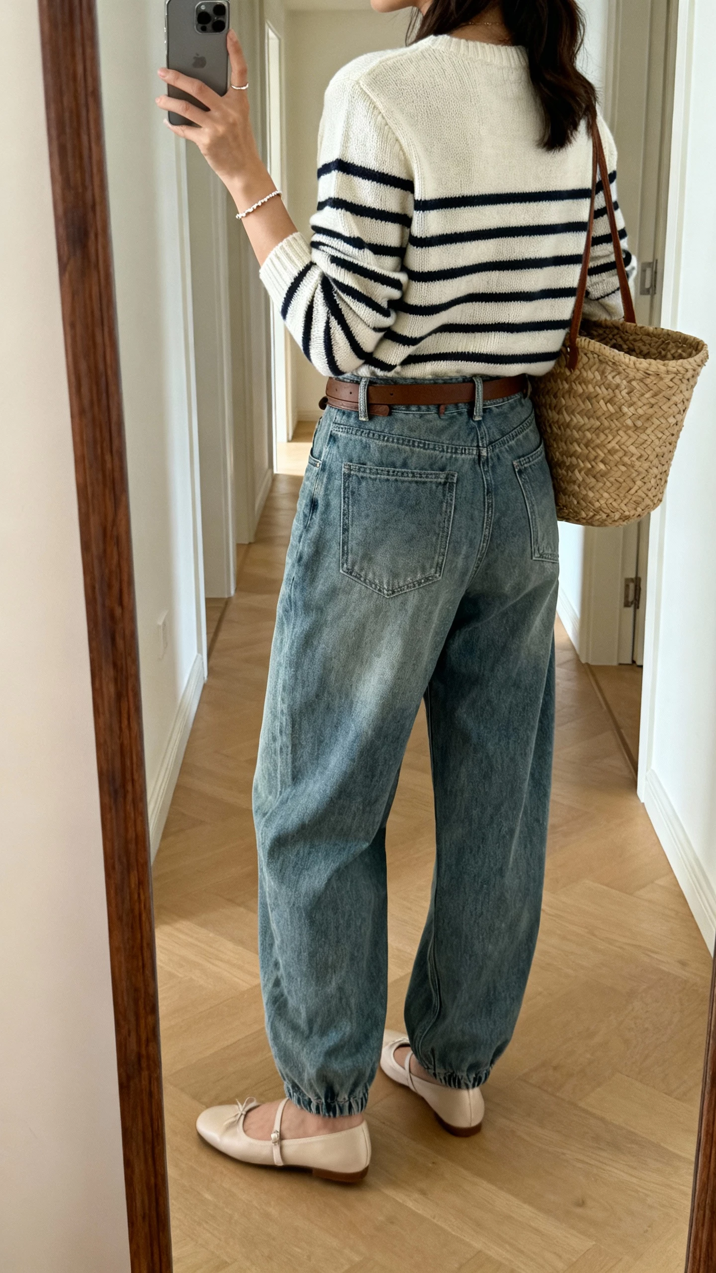 iPhone back-view selfie in a hallway mirror of a woman wearing a Parisian stripe knit, belted baggy jeans, ballet flats, basket bag, delicate bracelet; face not visible, morning light, natural iPhone quality.