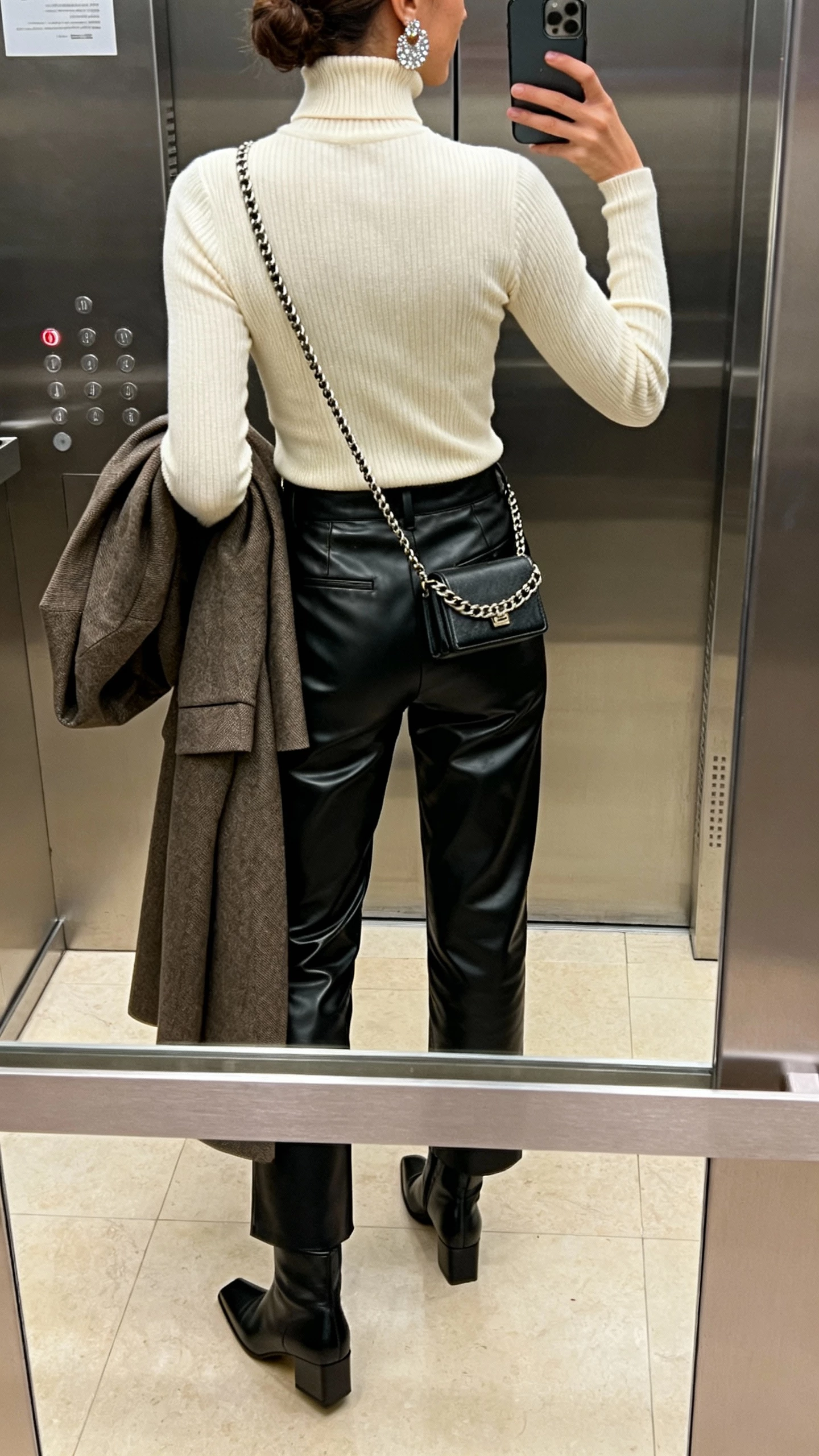 iPhone back-view selfie in an elevator mirror of a woman wearing straight-leg black leather pants, fitted cream ribbed turtleneck, square-toe ankle boots, sparkly statement earrings, compact chain-strap crossbody, tailored coat over one arm; face not visible, indoor lighting, natural iPhone photo quality.