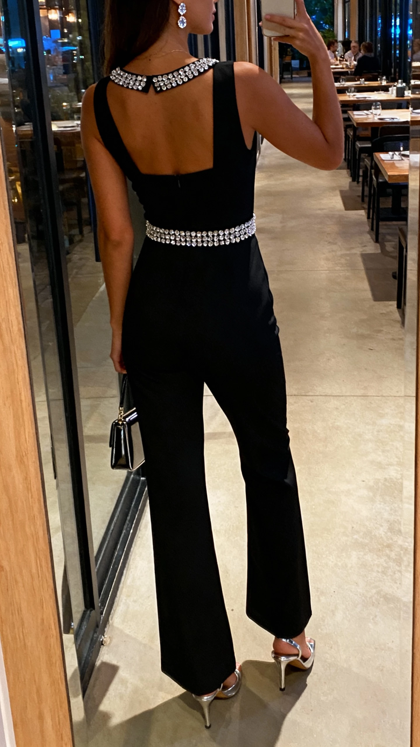 iPhone back-view selfie of a woman in a black statement jumpsuit with crystal accents at the neckline and waist, metallic heels, crystal stud earrings, and a sleek mini bag, face not visible, hallway mirror outside a restaurant, evening indoor lighting, natural iPhone photo quality.