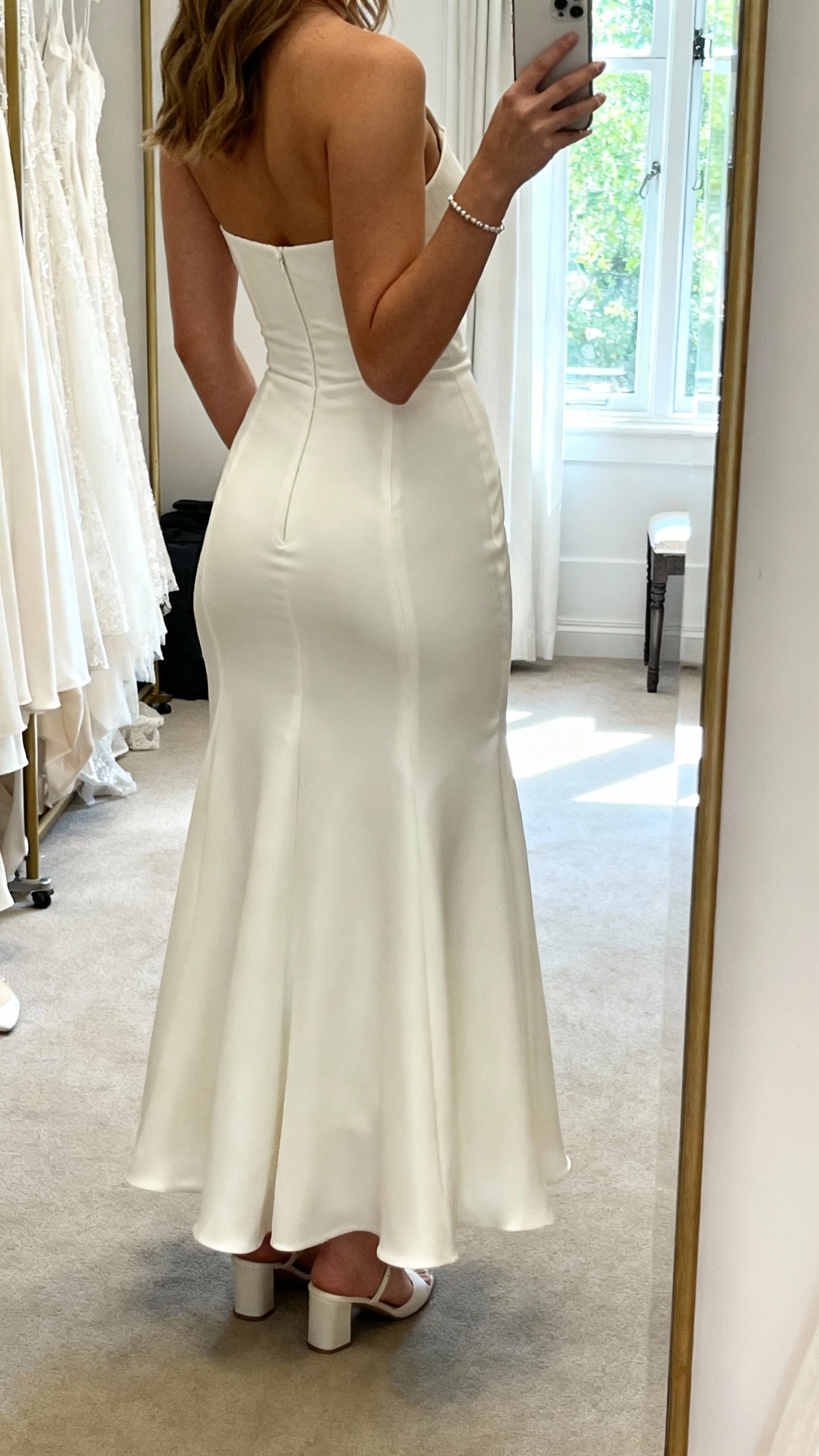 iPhone back-view selfie of a woman in a bridal gown with gentle hip shaping, smoothing side panels, and a softly skimming skirt over the seat; minimal bracelet and simple heels, face not visible, dressing room mirror, natural daylight, casual iPhone photo