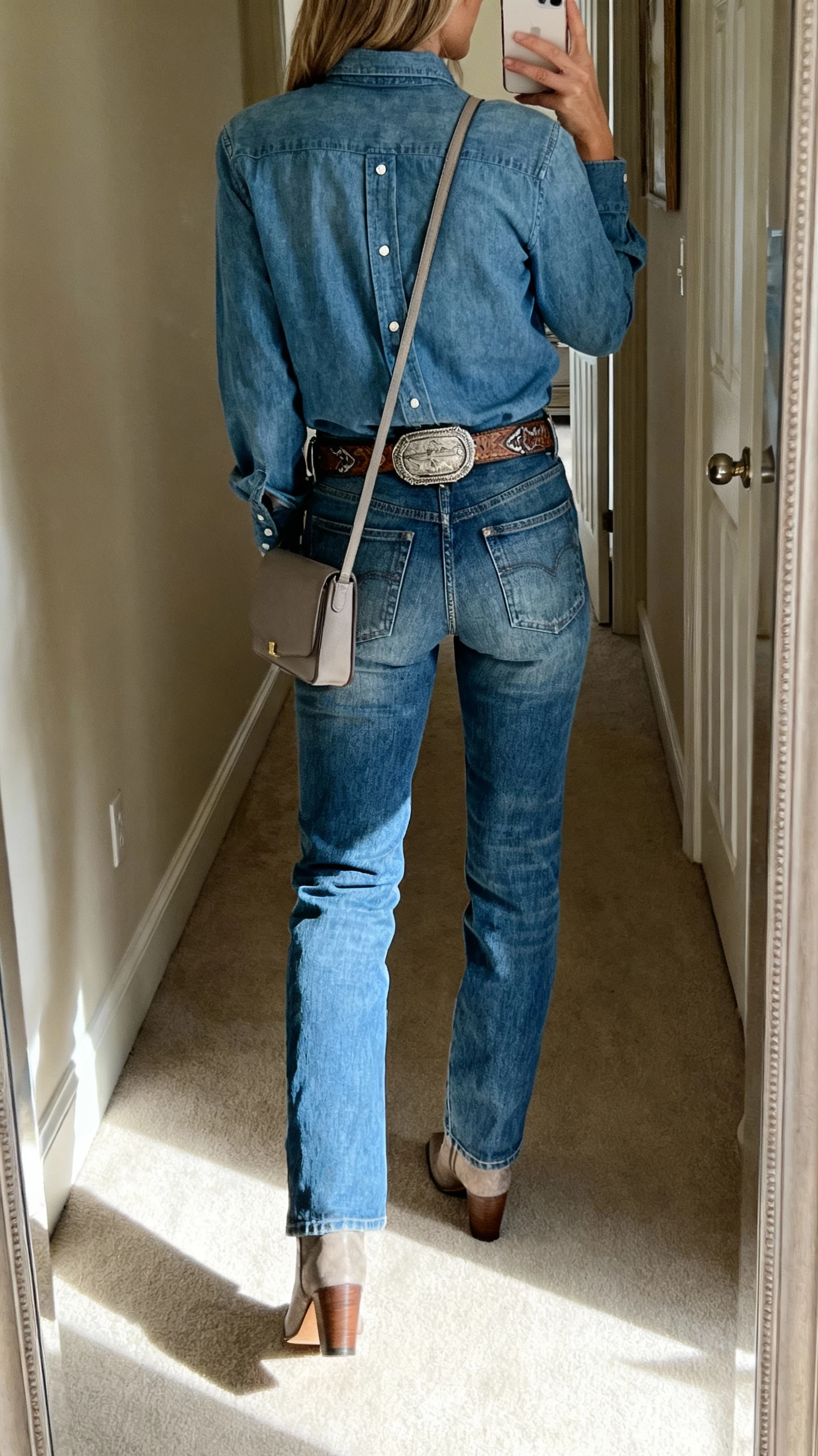 iPhone back-view selfie of a woman in a chambray button-down tucked into blue jeans in a similar wash, western-inspired belt, heeled ankle boots, minimal crossbody; hallway mirror, face not visible, morning light, natural iPhone photo quality.