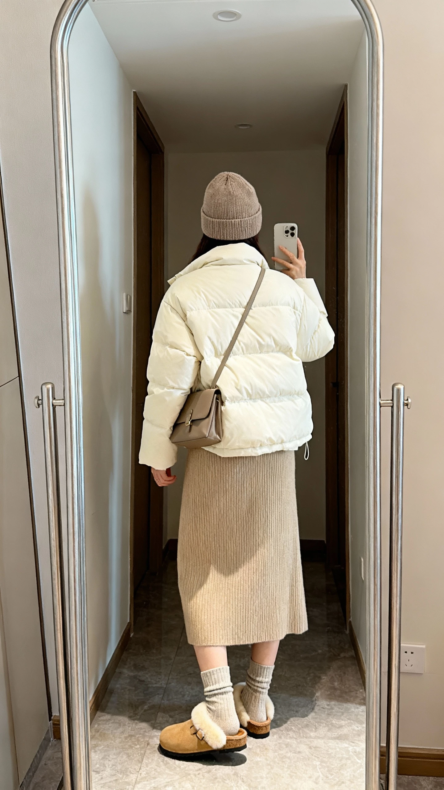 iPhone back-view selfie of a woman in a cream puffer jacket over a beige knit midi dress, cozy socks with tan shearling clogs, beanie, crossbody bag, face not visible, hallway mirror, soft indoor lighting, natural iPhone quality.
