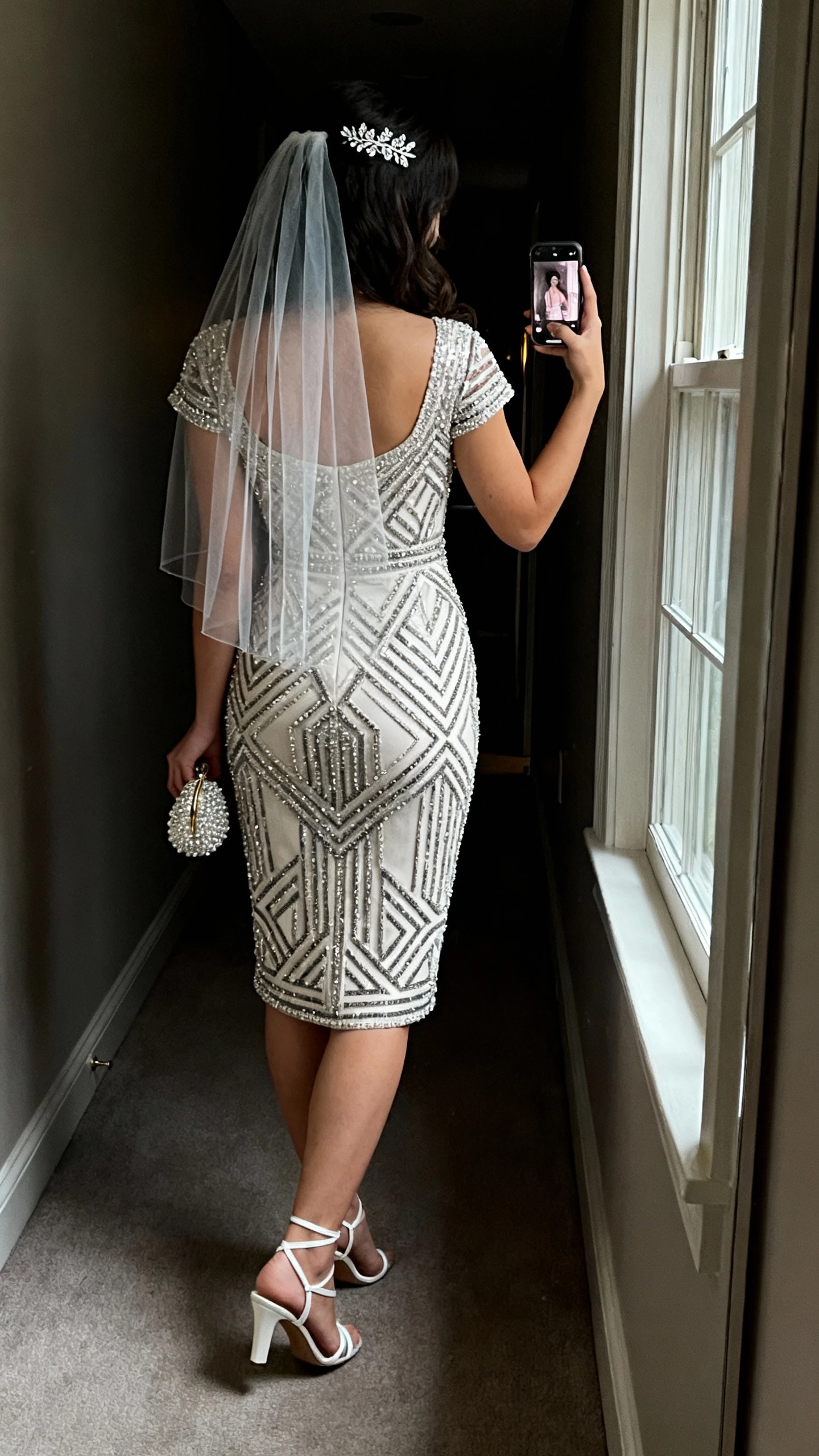 iPhone back-view selfie of a woman in a deco beaded sheath gown with geometric motifs and cap sleeves, art-deco T-strap heels, delicate crystal hair comb, sheer fingertip veil, small beaded clutch, face not visible, dim hallway near a window, evening indoor light, natural iPhone photo quality.