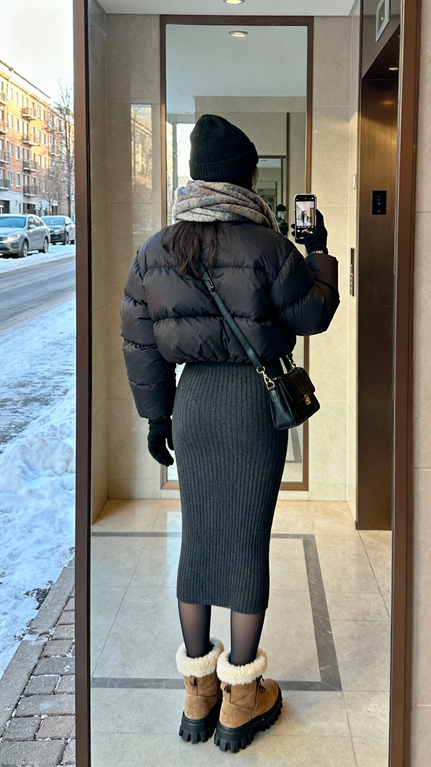 iPhone back-view selfie of a woman in a fitted ribbed knit midi dress, waist-length puffer, shearling-lined lug-sole boots, thermal tights, beanie, crossbody bag, cozy scarf and tech-friendly gloves, face not visible, apartment lobby mirror with snowy street visible, cool morning light, natural iPhone photo quality.