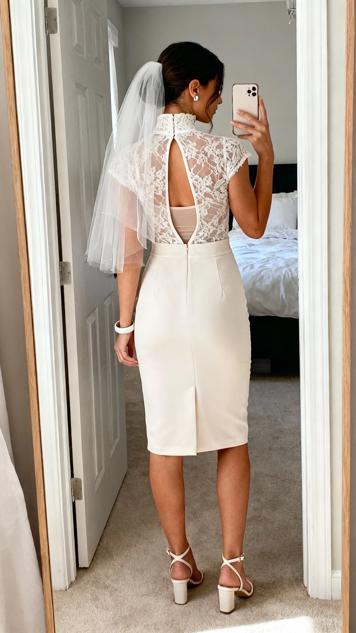 iPhone back-view selfie of a woman in a high-neck illusion lace bodice with a keyhole back, crepe sheath skirt, sleek fingertip veil, minimalist ankle-strap heels, modern stud earrings, cuff bracelet, seamless shapewear slip; bedroom doorway mirror, face not visible, soft morning light, natural iPhone photo quality.