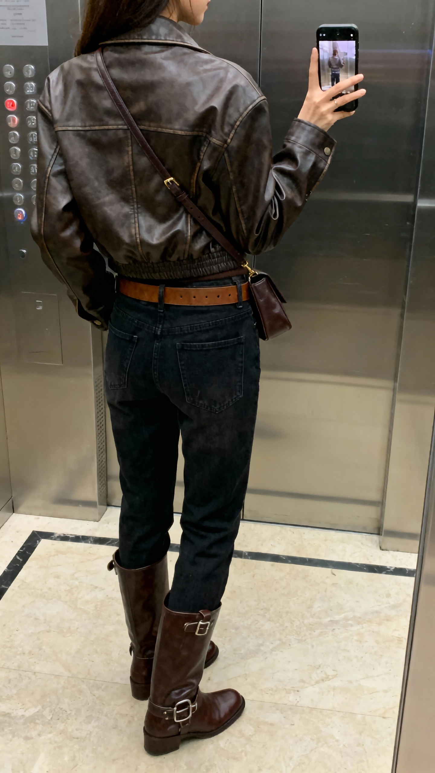iPhone back-view selfie of a woman in a leather jacket, matching-tone leather ankle boots and belt, dark jeans, tonal crossbody, face not visible, elevator mirror, indoor lighting, casual iPhone photo.