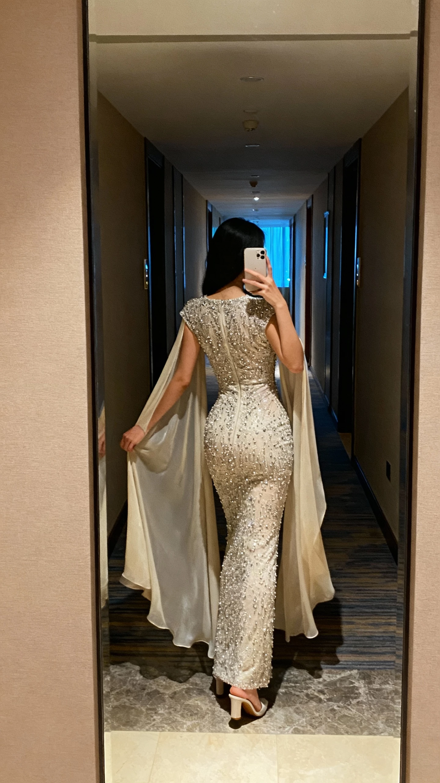 iPhone back-view selfie of a woman in a luxe beaded sheath gown with a flowing cape for effortless drama, minimal heels peeking out, face not visible, hotel corridor mirror, evening indoor lighting, natural iPhone photo quality