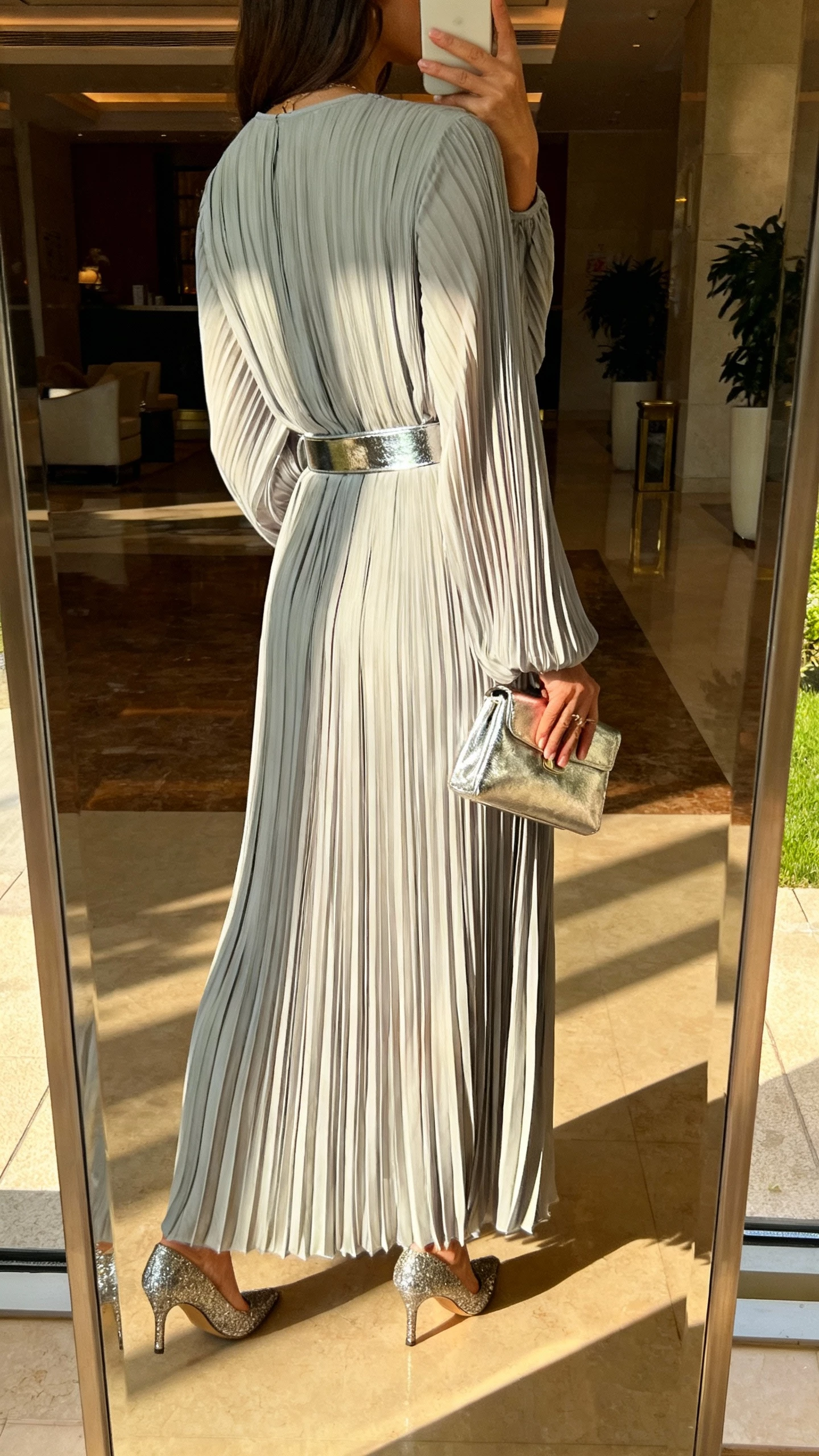 iPhone back-view selfie of a woman in a pleated maxi dress with metallic belt and metallic clutch, paired with shimmer heels, face not visible, taken in a lobby mirror, late afternoon daylight, casual iPhone vibe.