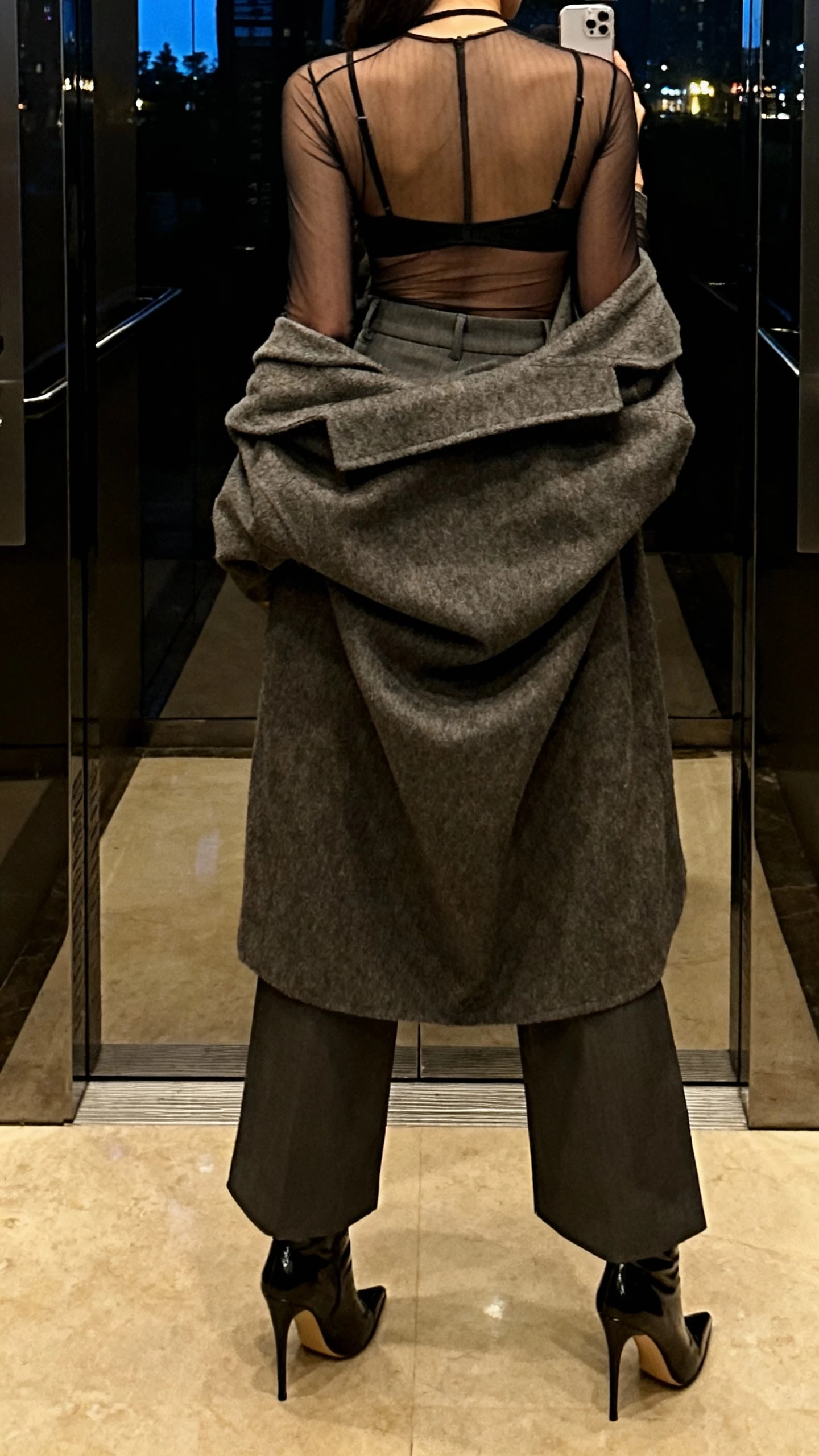 iPhone back-view selfie of a woman in a sheer mesh bodysuit layered over a black bralette, high-waisted wide-leg trousers, sleek stiletto booties, oversized wool coat thrown over shoulders, face not visible, elevator mirror with evening indoor lighting, natural iPhone photo.