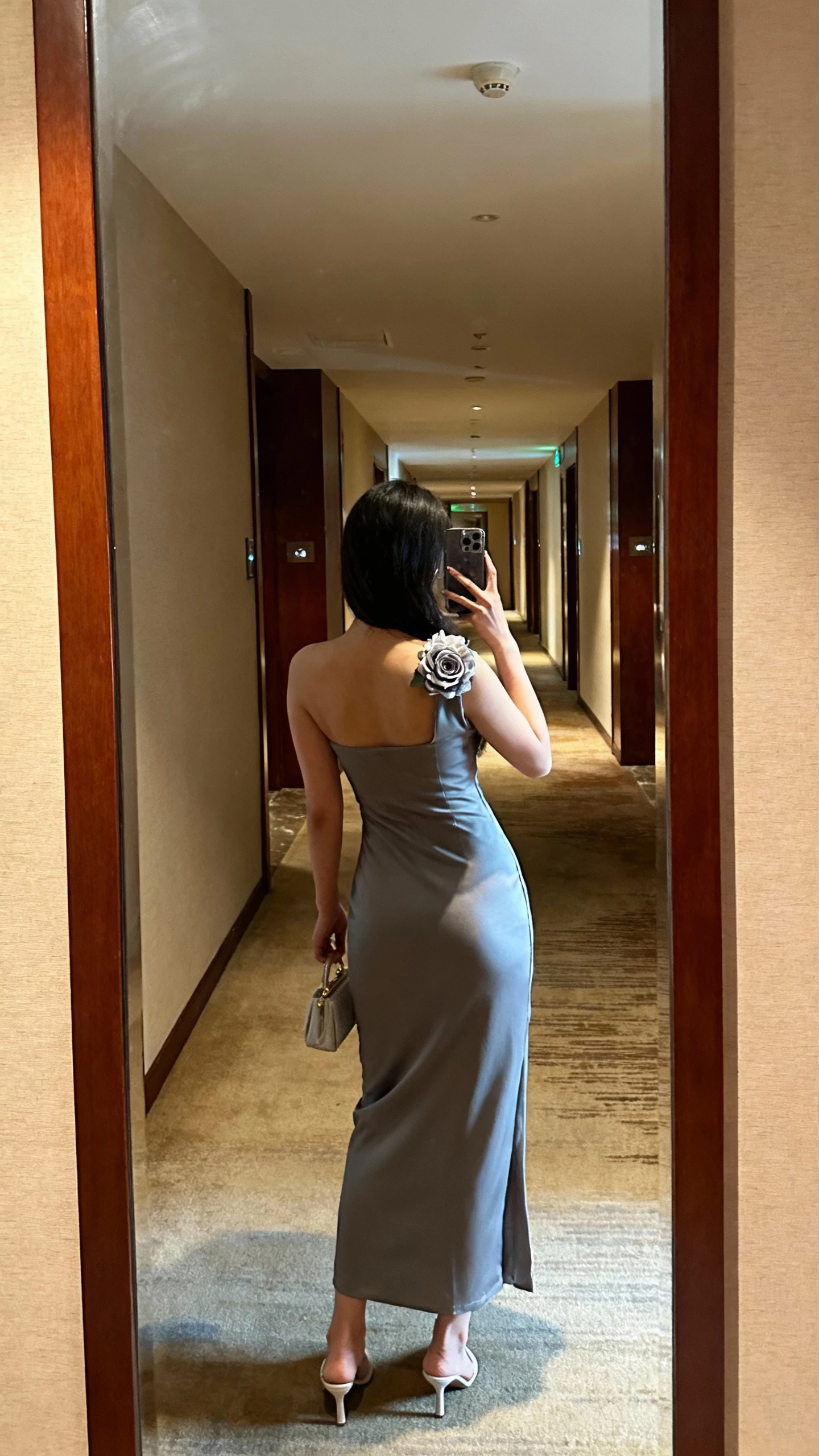 iPhone back-view selfie of a woman in a sleek column dress with a corsage detail at the shoulder, minimalist heels, small evening bag, face not visible, hotel corridor mirror before an event, warm indoor lighting, natural iPhone quality.