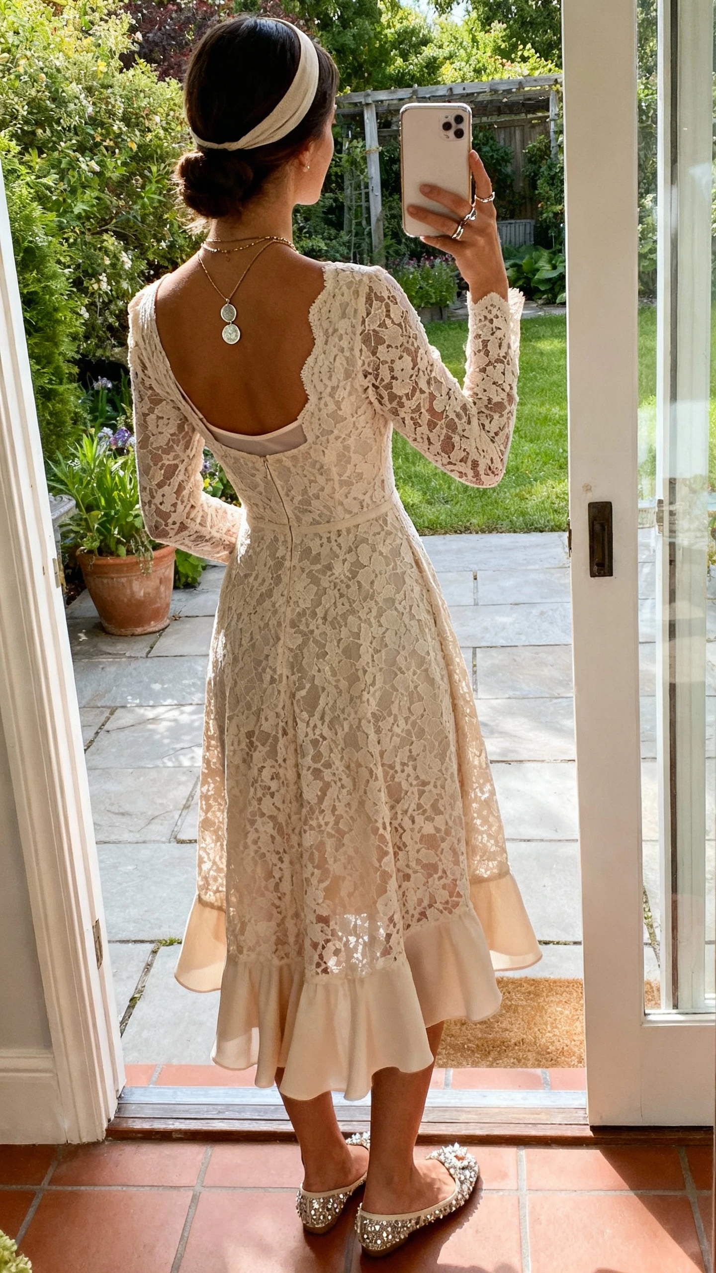 iPhone back-view selfie of a woman in a soft fit-and-flare guipure lace dress with long sleeves and a flutter train, no veil with a minimalist headband, embellished flats, layered coin necklace and delicate rings, seamless slip; garden patio doorway, face not visible, afternoon natural light, natural iPhone photo quality.