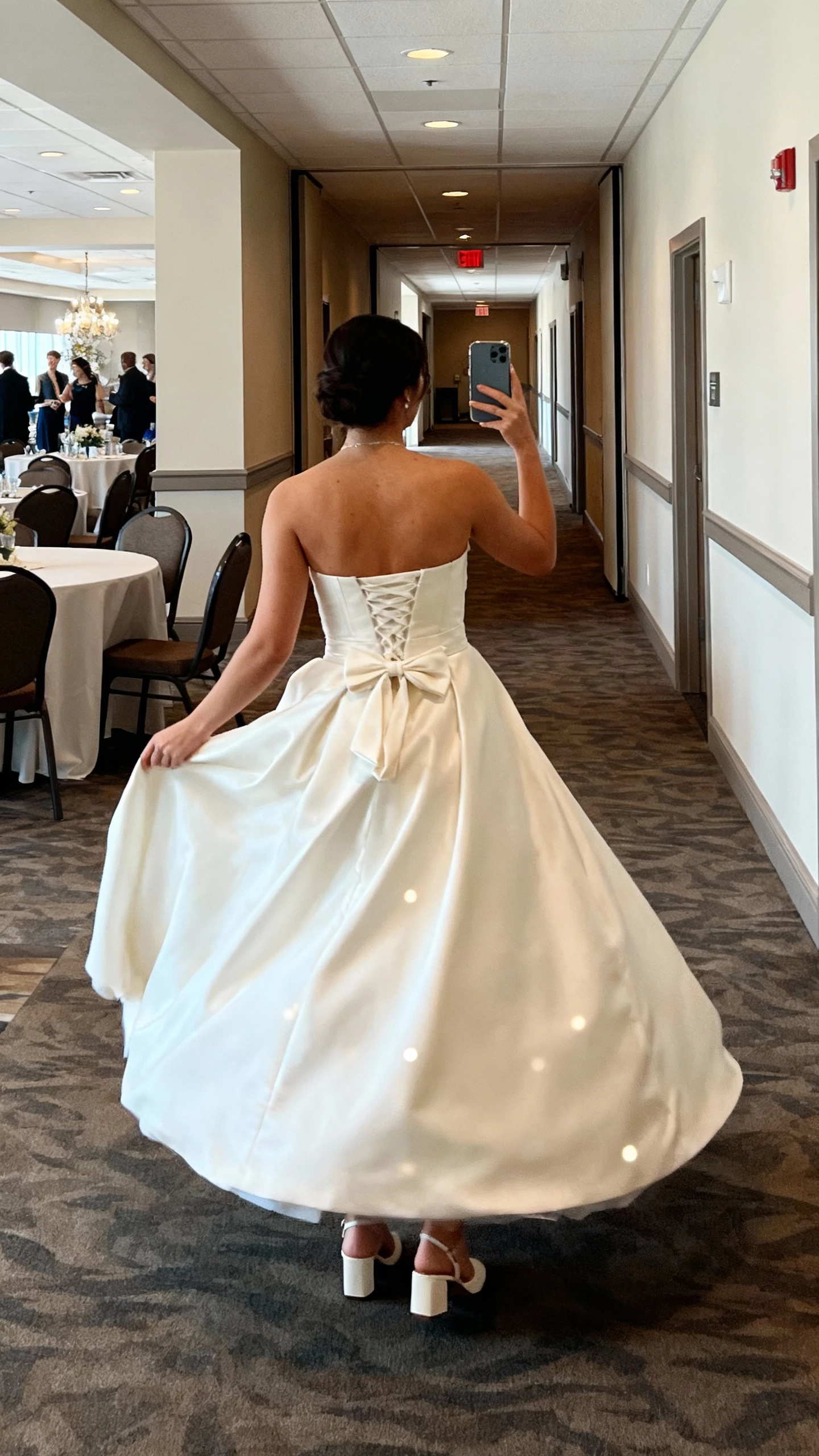 iPhone back-view selfie of a woman in a wedding gown with a custom bustle and mapped movement points so the skirt lifts neatly for dancing; comfortable block heels, face not visible, reception hall corridor, ambient indoor lighting, natural iPhone photo quality