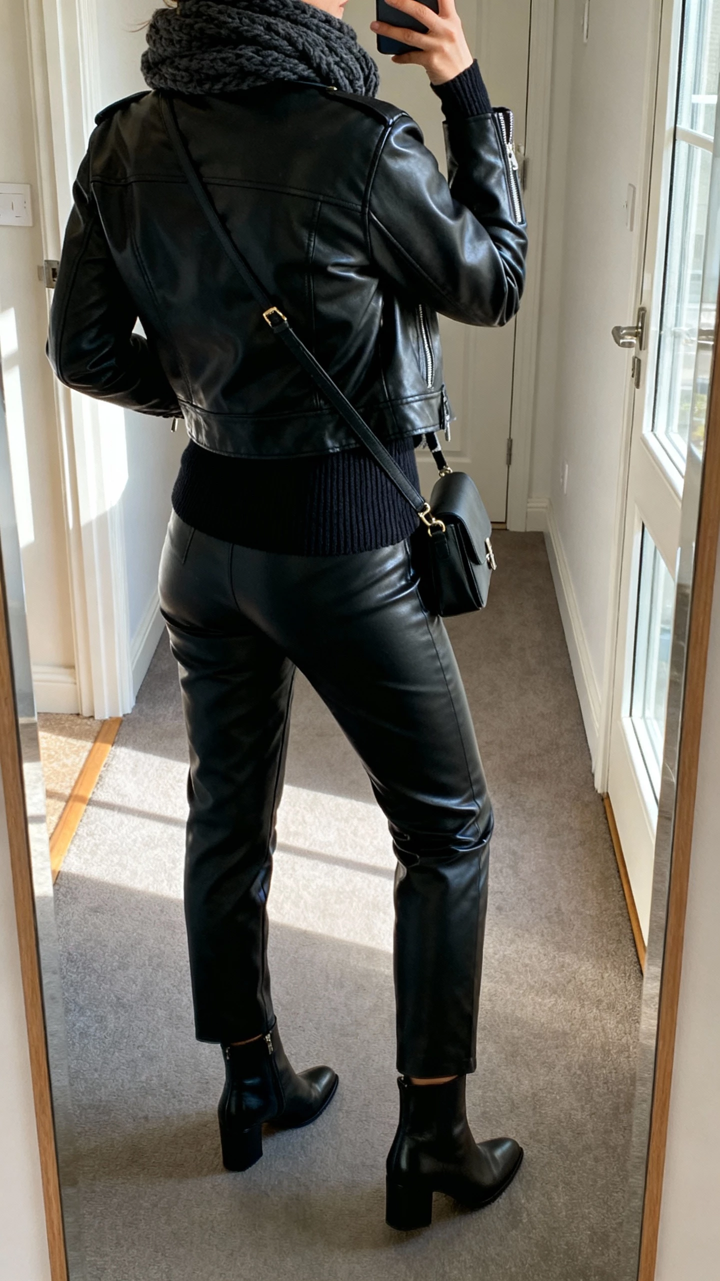 iPhone back-view selfie of a woman in an all-black leather look: leather jacket, leather pants, fitted knit underneath, chunky scarf, sleek ankle boots, and a crossbody bag, face not visible, hallway mirror by apartment door, natural daylight from window, casual iPhone photo.