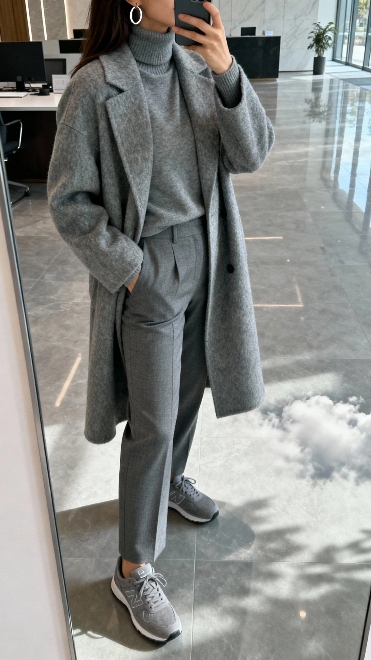 iPhone back-view selfie of a woman in an all-gray outfit: gray wool coat, gray turtleneck, gray tailored trousers, gray sneakers, silver hoop earrings, face not visible, office lobby mirror, cloudy daylight, natural iPhone photo quality