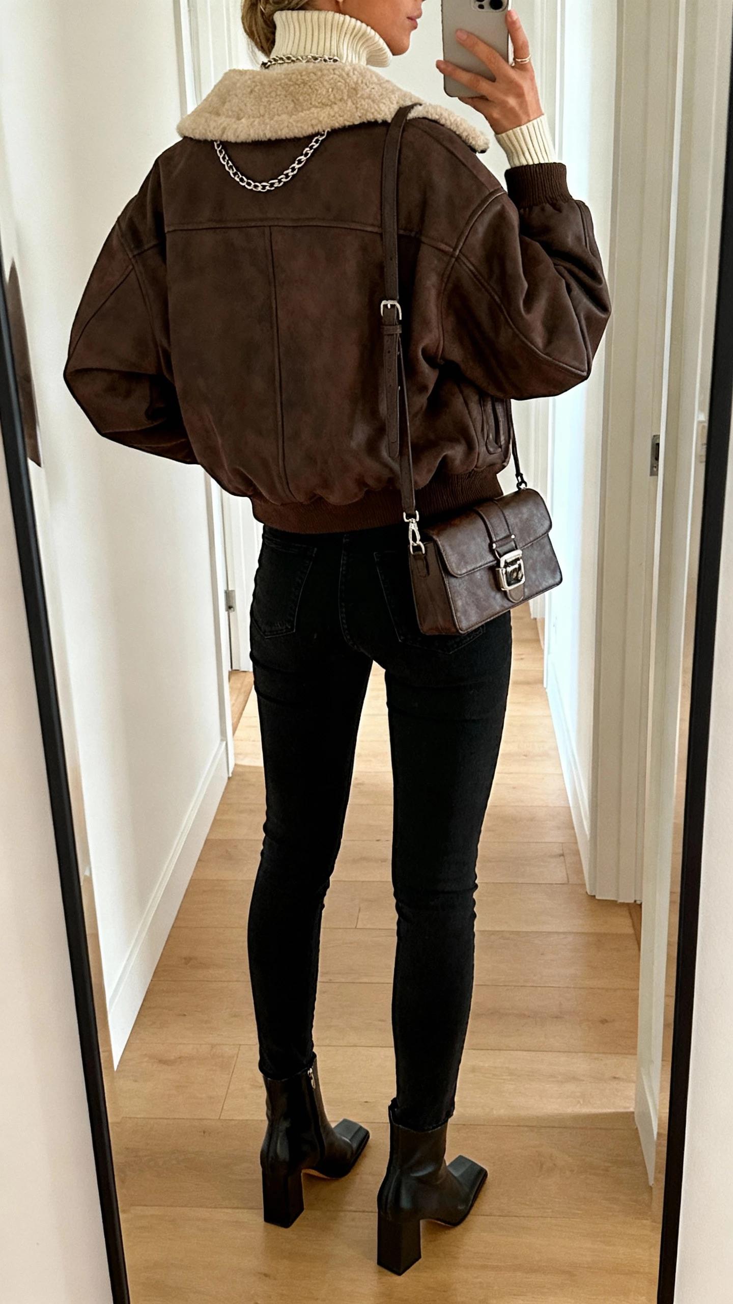 iPhone back-view selfie of a woman in an oversized espresso shearling-lined aviator jacket, cream ribbed mock-neck top, high-waist black skinny jeans, square-toe heeled ankle boots, structured crossbody with metal hardware, delicate chain necklace peeking; hallway mirror, face not visible, warm indoor lighting, natural iPhone photo.