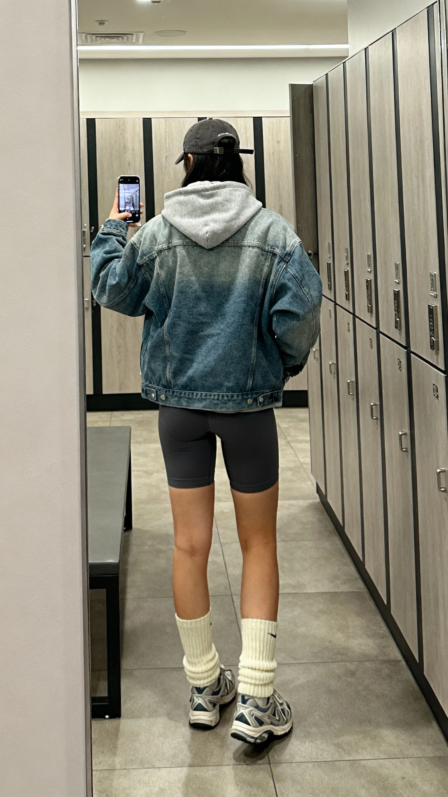 iPhone back-view selfie of a woman in an oversized jean jacket layered over a hoodie, bike shorts, high crew socks, sporty trainers, baseball cap tucked low, face not visible, gym locker room mirror, soft indoor lighting, casual iPhone photo
