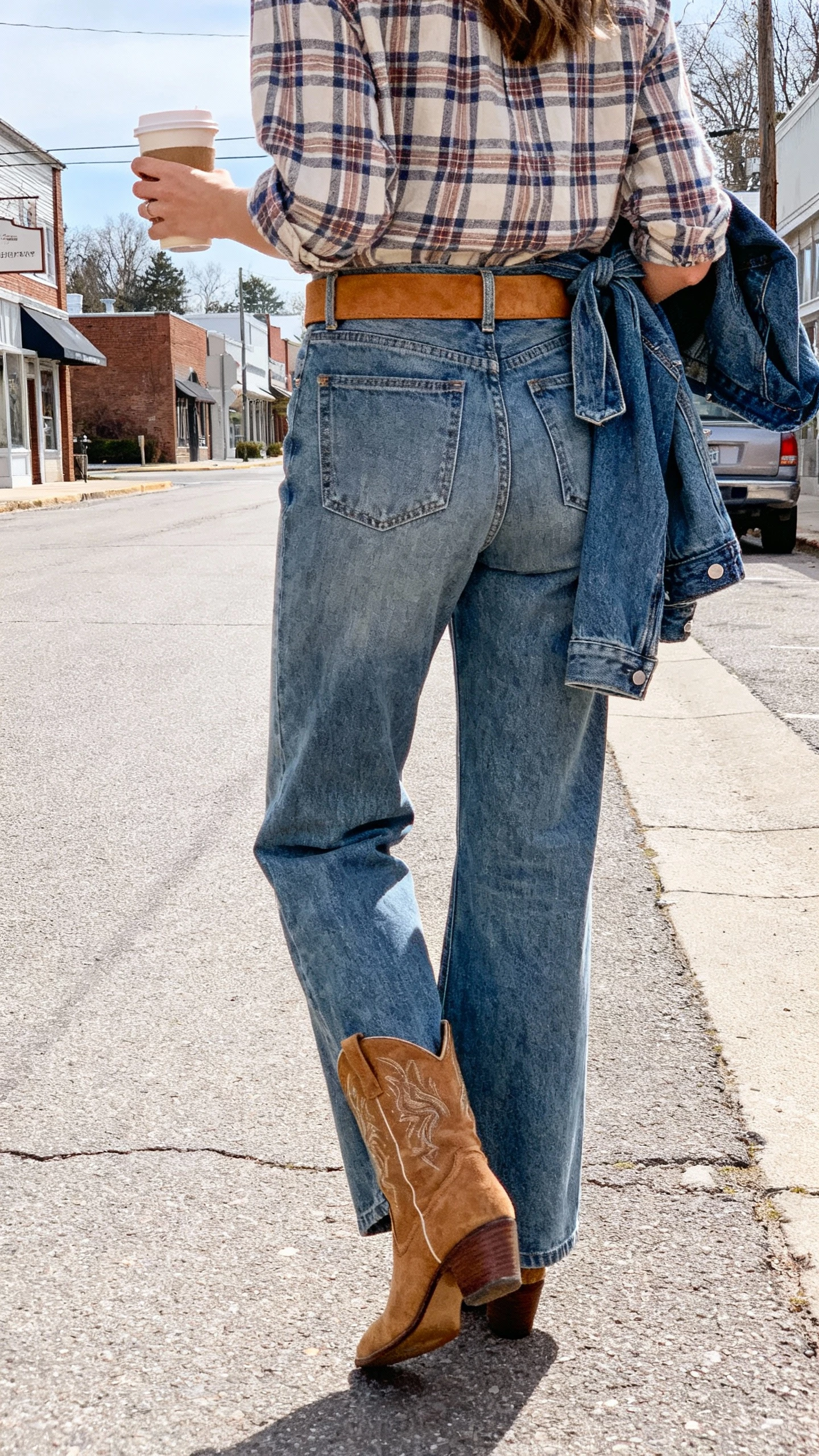 iPhone back-view selfie of a woman in medium-wash bootcut jeans with a tucked-in plaid shirt, a suede tan belt, low-heel western ankle boots, and a denim jacket tied at the waist, holding a coffee; face not visible, small-town street in daylight, casual iPhone photo.
