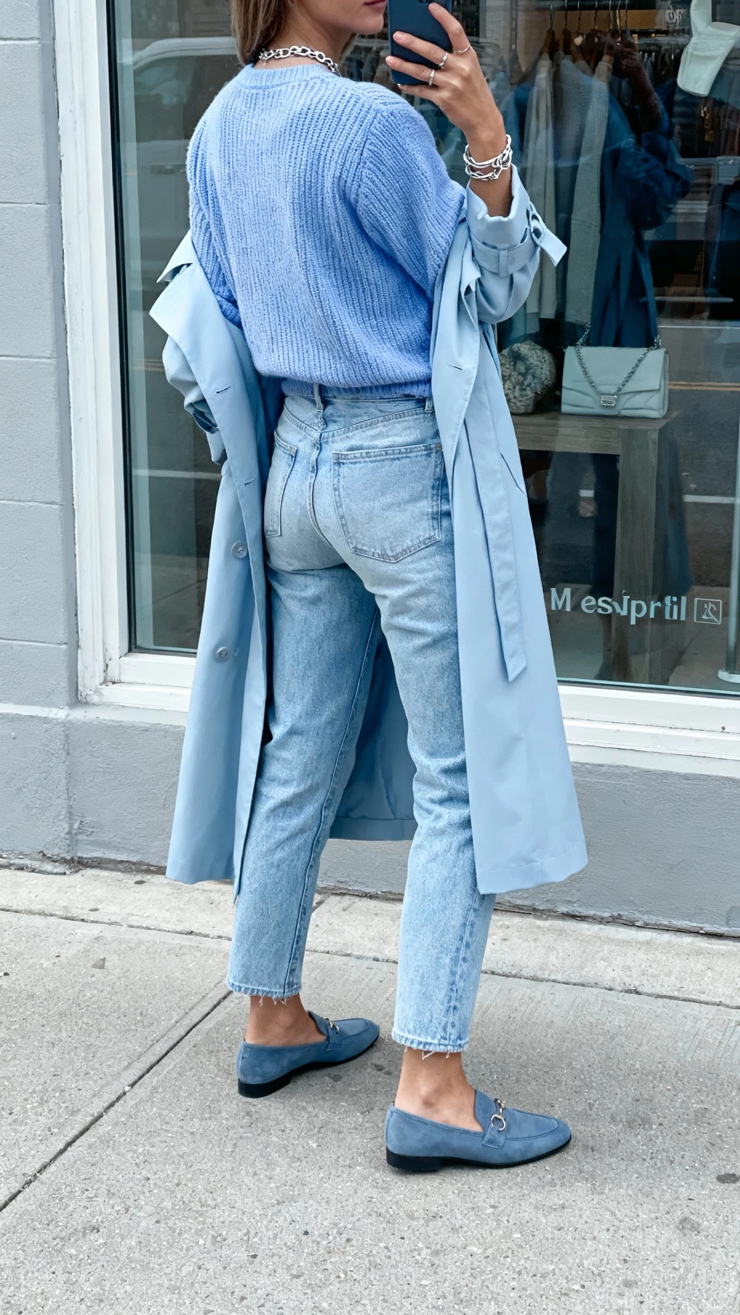 iPhone back-view selfie of a woman in monochrome soft blues: powder-blue knit, light blue jeans, pale blue trench, slate-blue loafers, silver jewelry, face not visible, street reflection in a shop window, overcast natural light, natural iPhone aesthetic