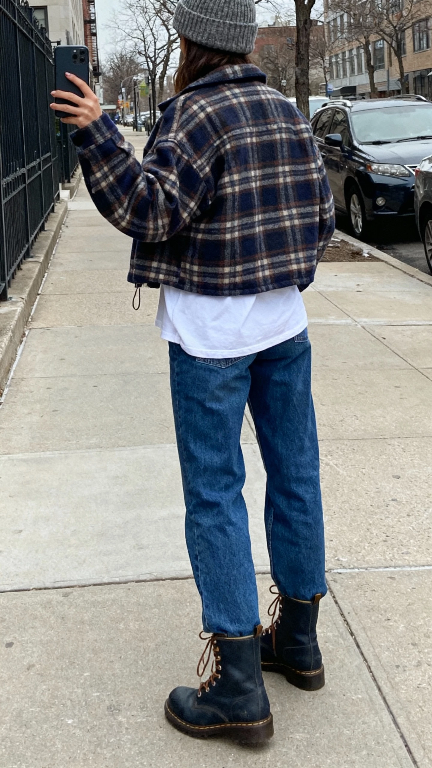 iPhone back-view selfie of a woman layering a plaid shacket over a white tee with straight blue jeans, lace-up boots, knit beanie, face not visible, city sidewalk, overcast natural light, iPhone candid feel