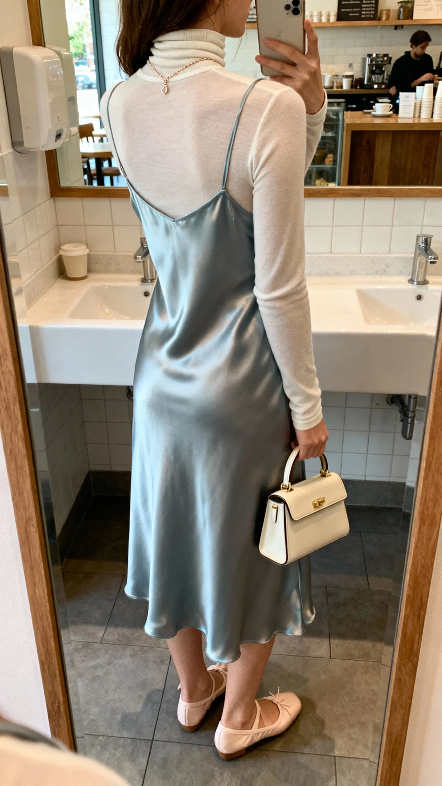 iPhone back-view selfie of a woman layering a thin turtleneck under a satin slip dress, with ballet flats, mini top-handle bag, dainty necklace, face not visible, cafe restroom mirror, soft morning light, natural iPhone photo.