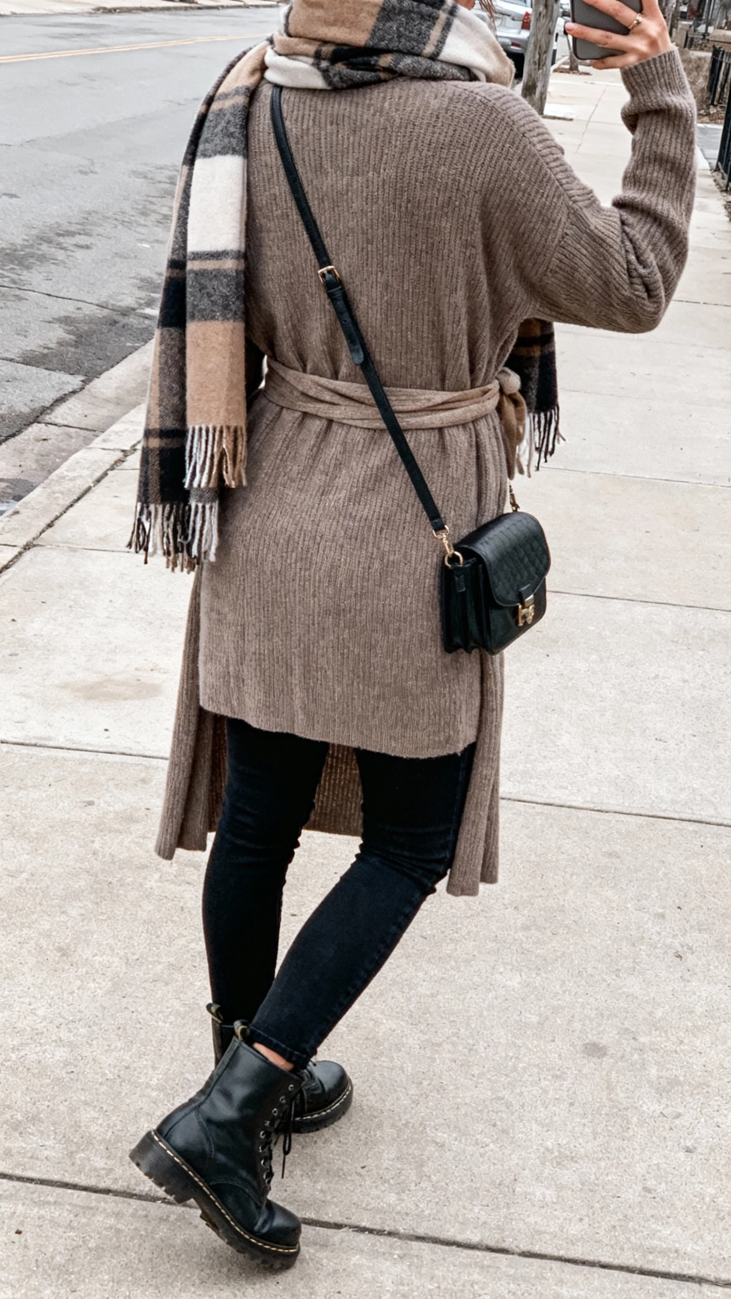 iPhone back-view selfie of a woman wearing a belted blanket scarf over a long cardigan, black jeans, and combat boots, crossbody bag on hip, face not visible, urban sidewalk setting with overcast daylight, natural iPhone photo quality.
