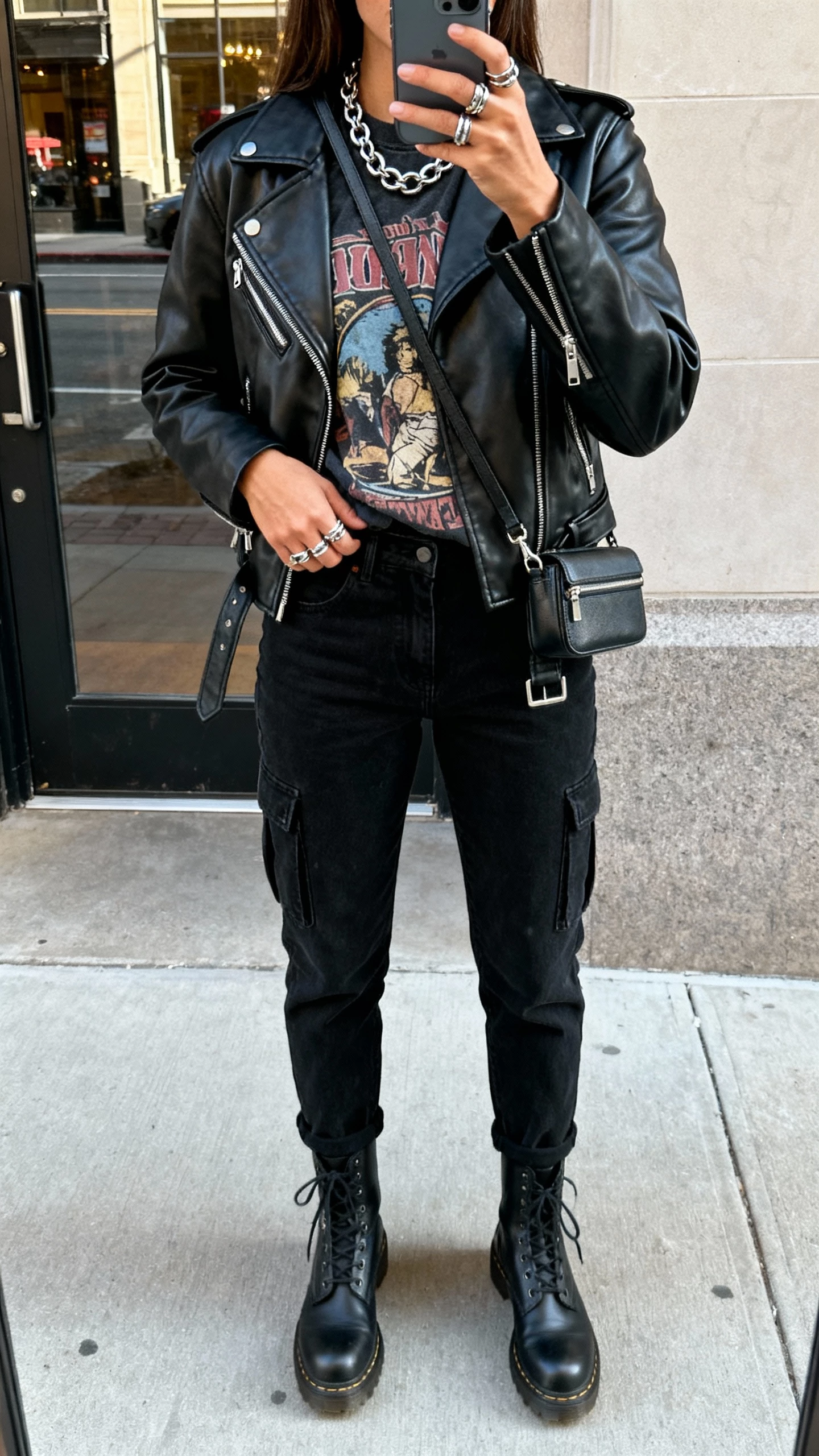 iPhone back-view selfie of a woman wearing a black leather moto jacket over a vintage graphic tee, black tapered cargo jeans, combat boots, chunky chain necklace, stacked rings, and a mini crossbody, face not visible, city street doorway reflection, natural daylight, casual iPhone photo.
