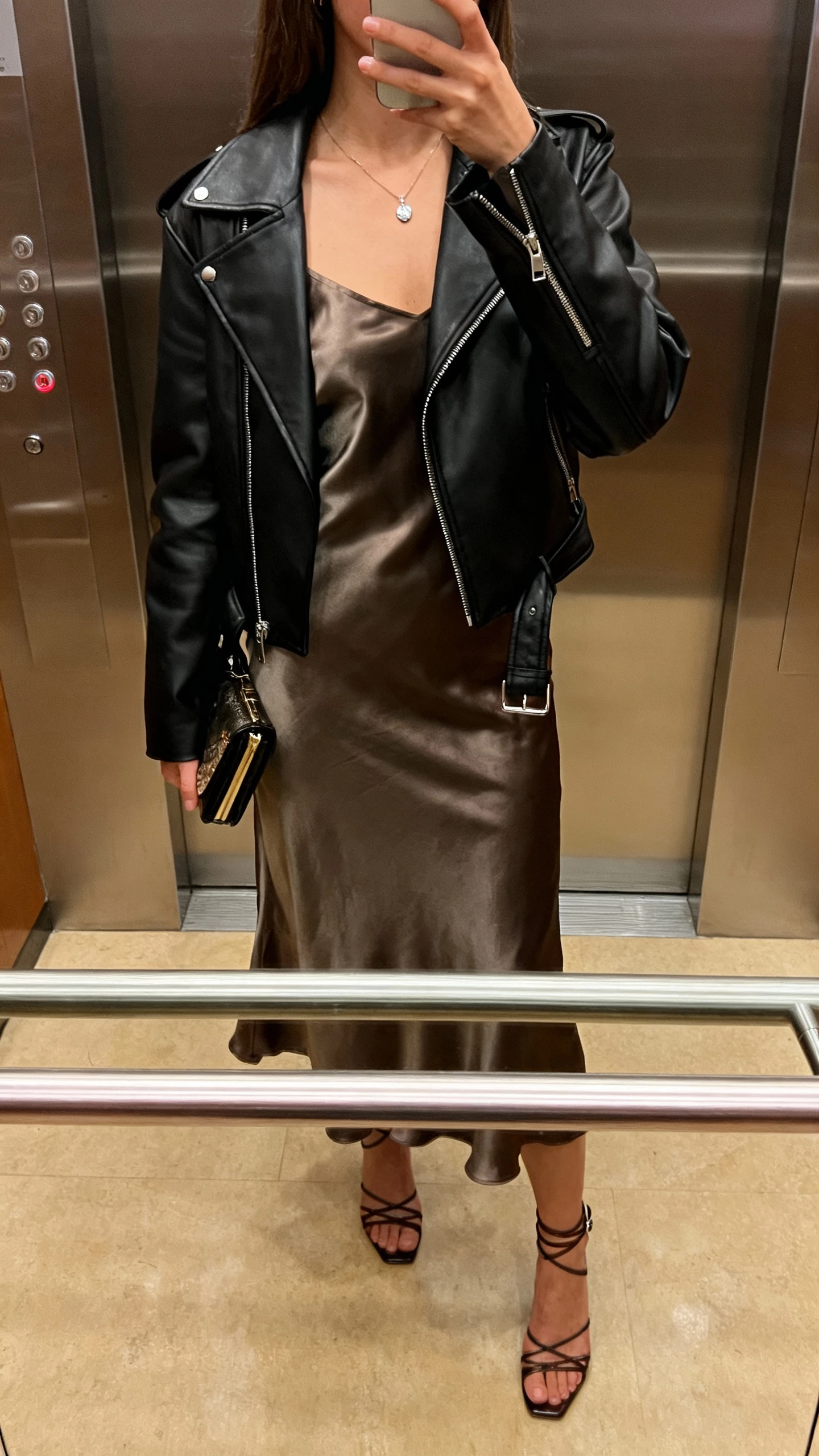 iPhone back-view selfie of a woman wearing a black leather jacket over a satin slip dress, delicate pendant necklace, strappy heels, clutch, face not visible, taken in an elevator mirror, warm indoor lighting, natural iPhone aesthetic.