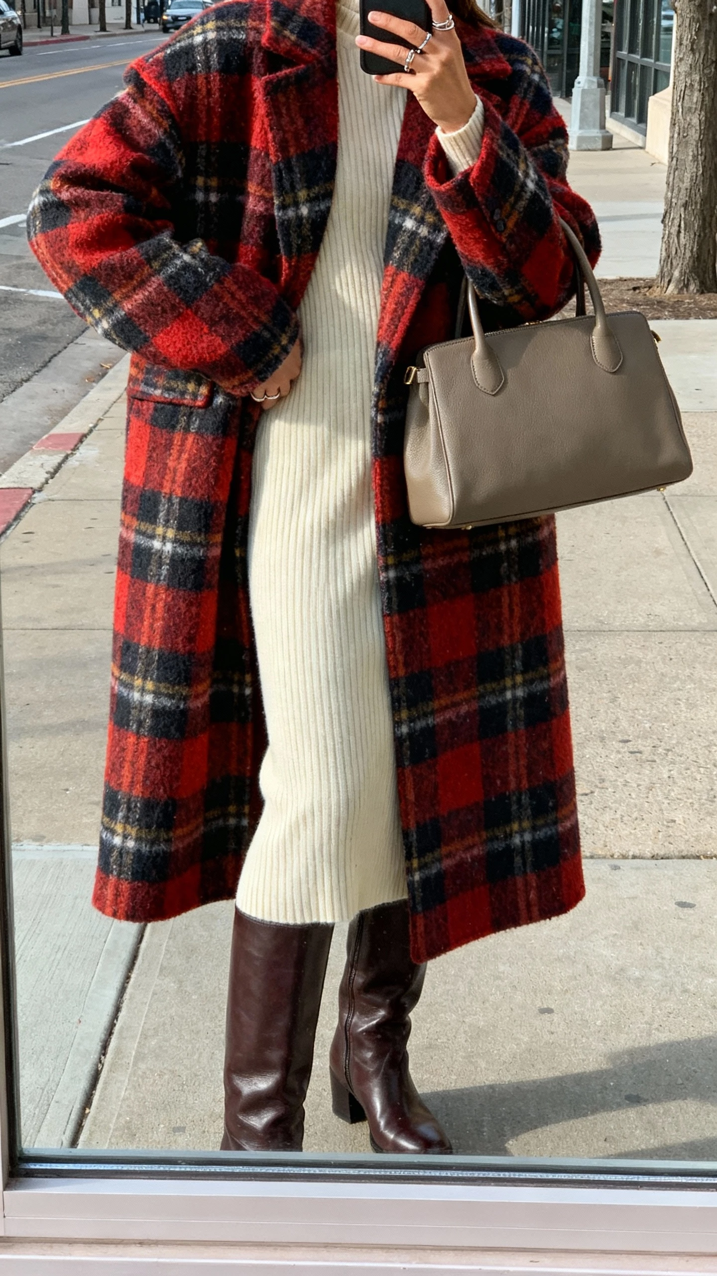 iPhone back-view selfie of a woman wearing a bold plaid statement coat over a ribbed cream knit midi dress, tall leather boots, structured top-handle bag, delicate rings, face not visible, captured on a city sidewalk window reflection, natural afternoon light, natural iPhone photo quality