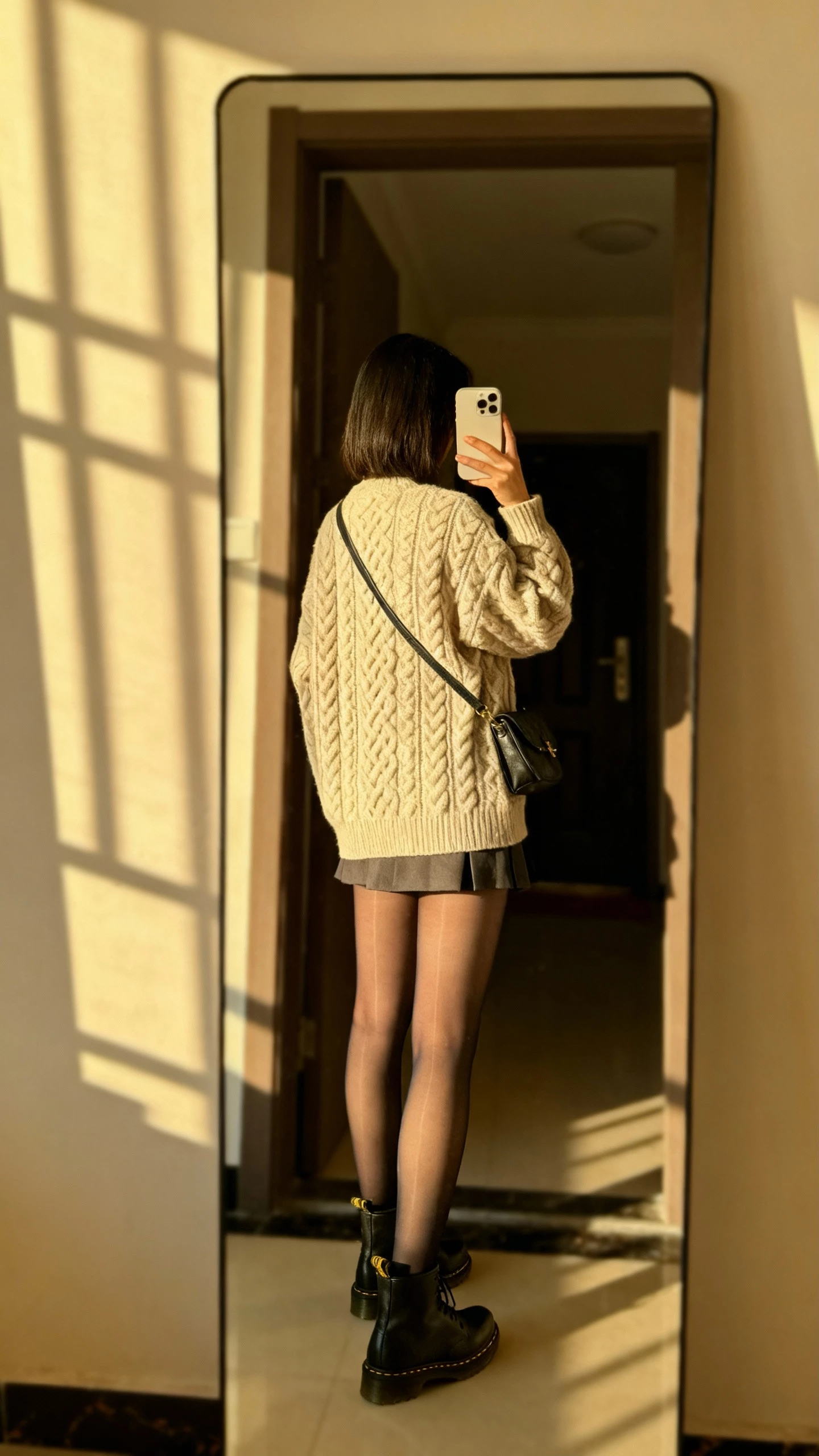 iPhone back-view selfie of a woman wearing a cable knit sweater, mini skirt, sheer tights, Doc Martens, crossbody bag, face not visible, apartment entry mirror, afternoon window light, natural iPhone photo.