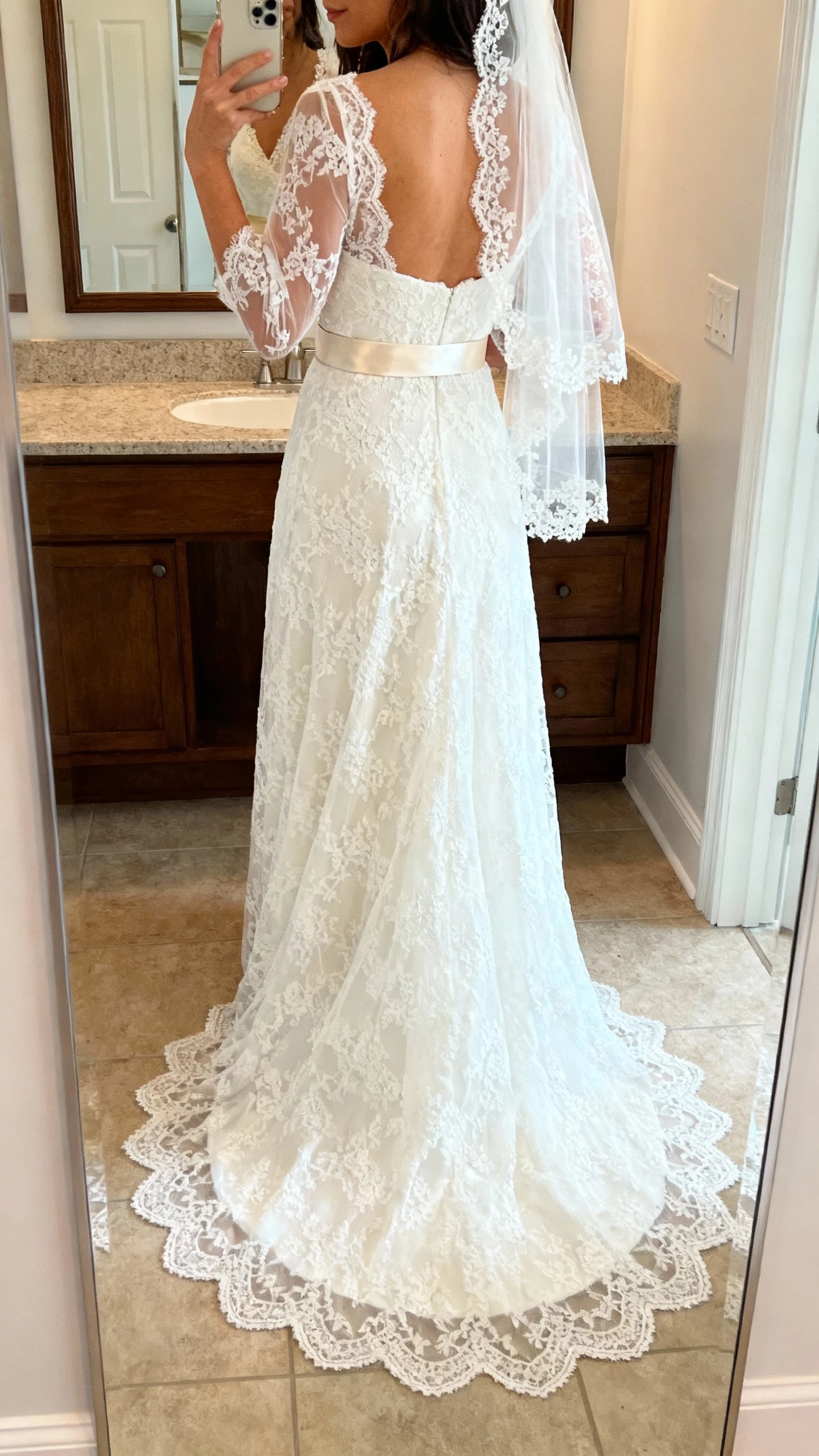 iPhone back-view selfie of a woman wearing a classic Chantilly lace wedding dress with sheer lace sleeves, scalloped lace train, satin ribbon belt, small lace-edged veil, face not visible, bathroom mirror, soft indoor lighting, natural iPhone photo quality