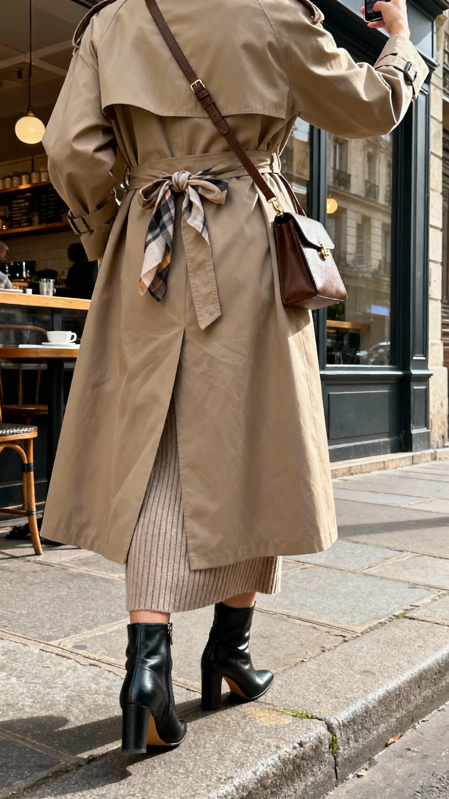 iPhone back-view selfie of a woman wearing a classic trench coat over a ribbed knit dress, heeled boots, crossbody bag, scarf tied to strap, face not visible, city sidewalk near café, soft morning daylight, natural iPhone photo quality