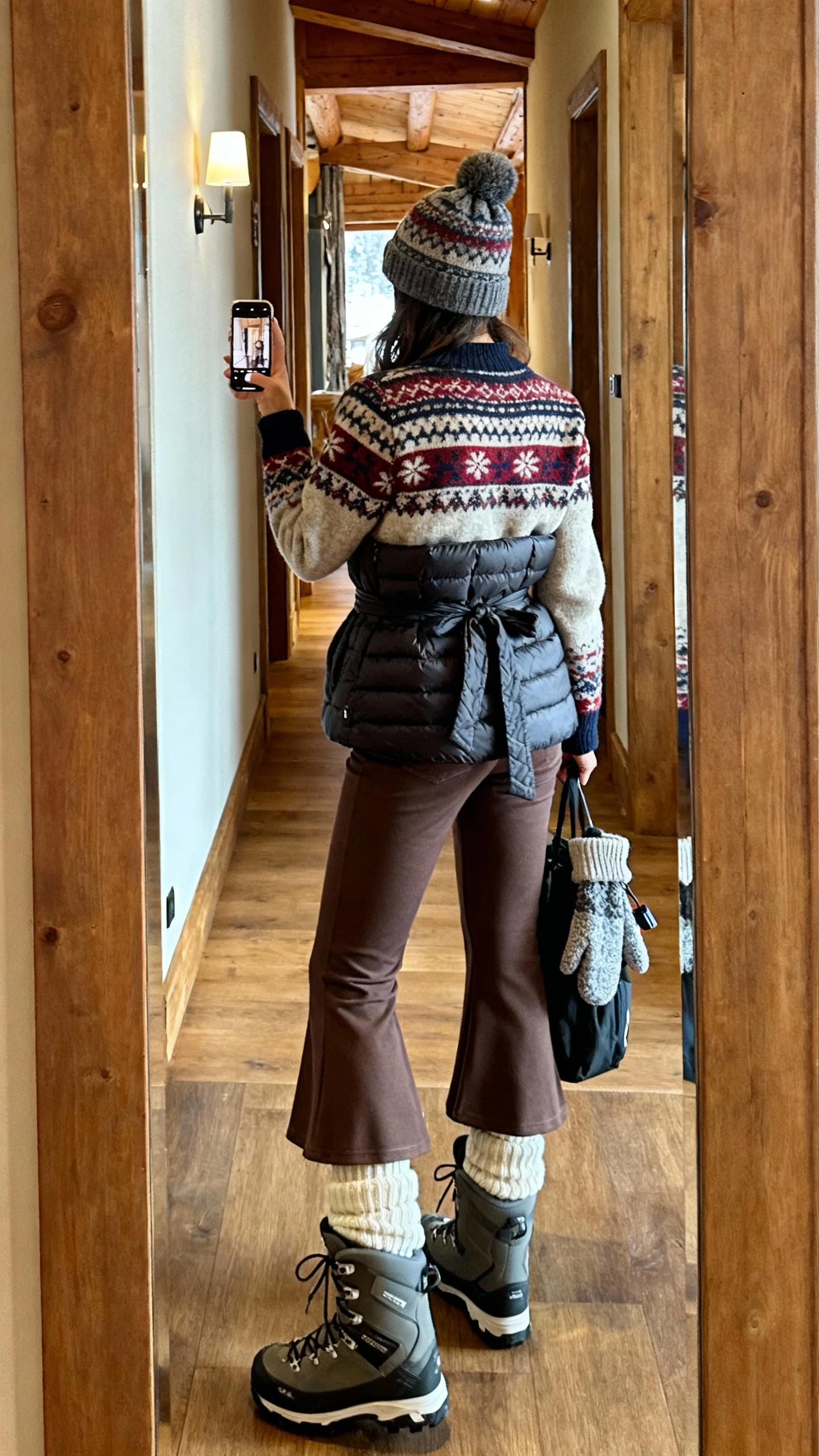 iPhone back-view selfie of a woman wearing a Fair Isle sweater, high-waist flared pants, quilted vest tied at the waist, thick wool socks peeking from snow-ready lace-up boots, knit beanie and mittens clipped to bag, face not visible, ski-lodge hallway mirror with warm indoor lighting, casual iPhone photo